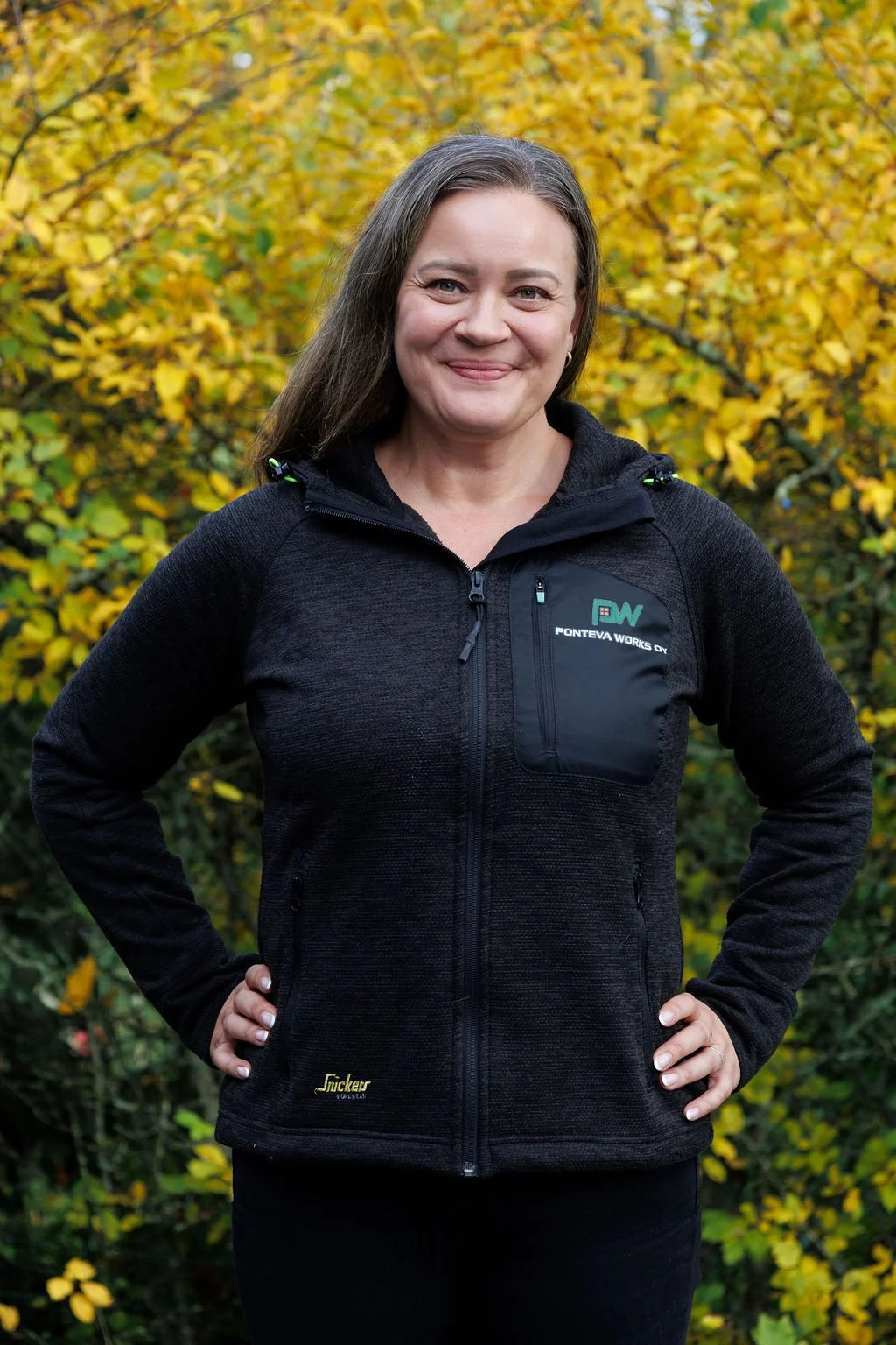 Ponteva Works Minna hymyilee ulkona syksyllä, taustalla keltaisia lehtiä, hänellä on päällä musta ulkoilupuku ja kädet lanteillaan.