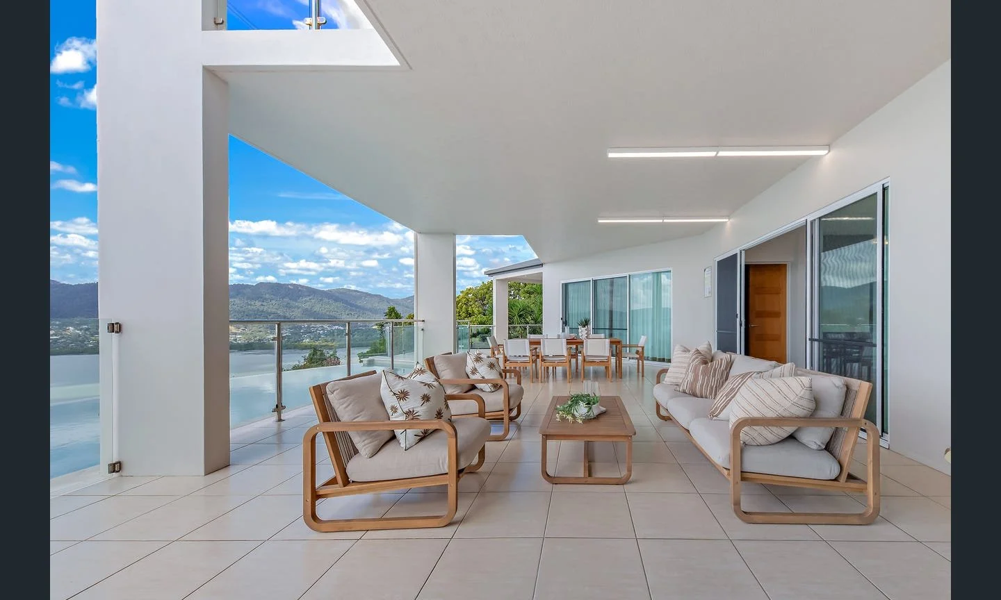 Modern balcony with outdoor seating, including three couches with cushions, a coffee table with a plant, a dining table with chairs, overlooking a lake and mountains in the background.