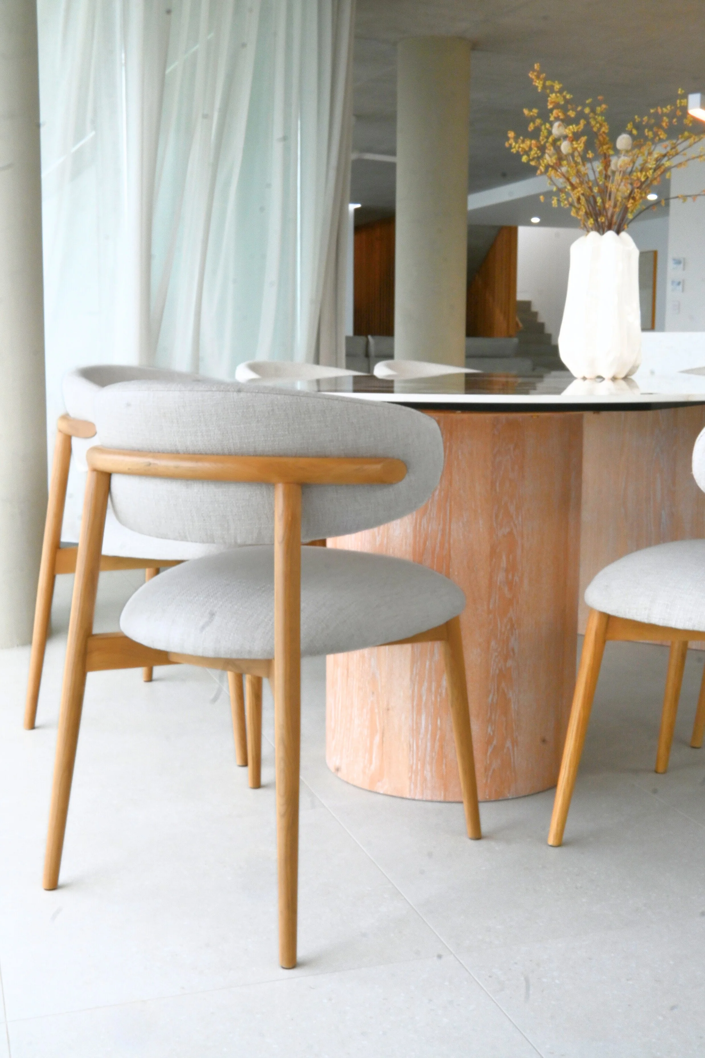 A modern dining area with a round wooden table and four beige upholstered chairs with wooden legs. A white vase with autumn-themed flowers sits on the table. In the background, there are light-colored curtains, a concrete pillar, and a staircase with