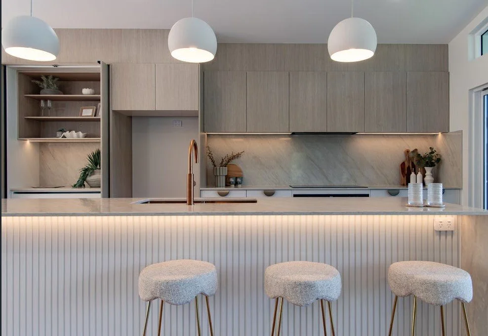 Modern kitchen with light wood cabinetry, marble backsplash, white countertop island with beaded detail, bang-graphite faucet, three beige upholstered bar stools, pendant lighting, open shelving with decorative items, and large window.