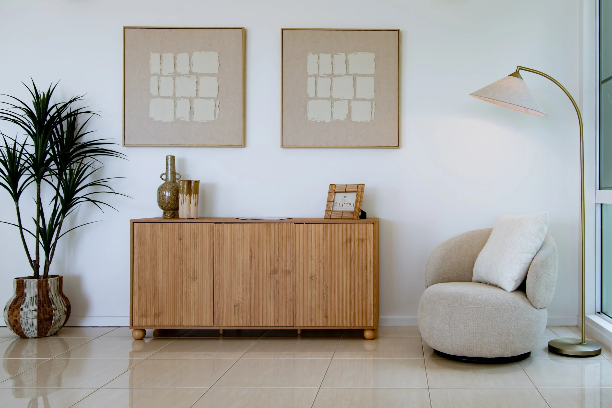 Modern interior living room with a wooden sideboard, abstract art on the walls, a large potted plant, a beige armchair, and a gold floor lamp.