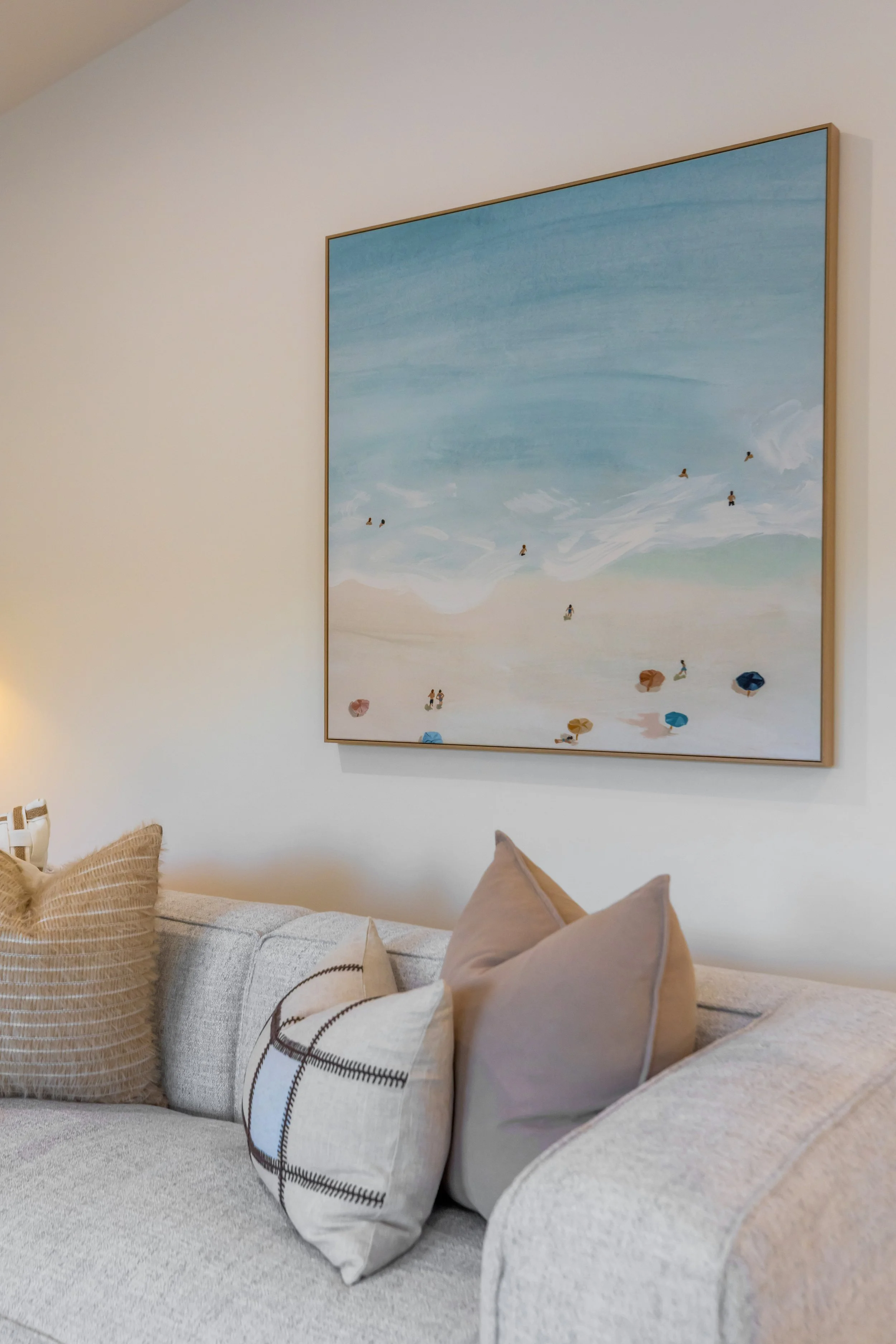 A cozy living room with a beige sofa decorated with various pillows, including a large beige pillow, a plaid pillow, and a taupe pillow. On the wall behind the sofa, there is a large painting of a beach scene with blue water and sand, and small beach