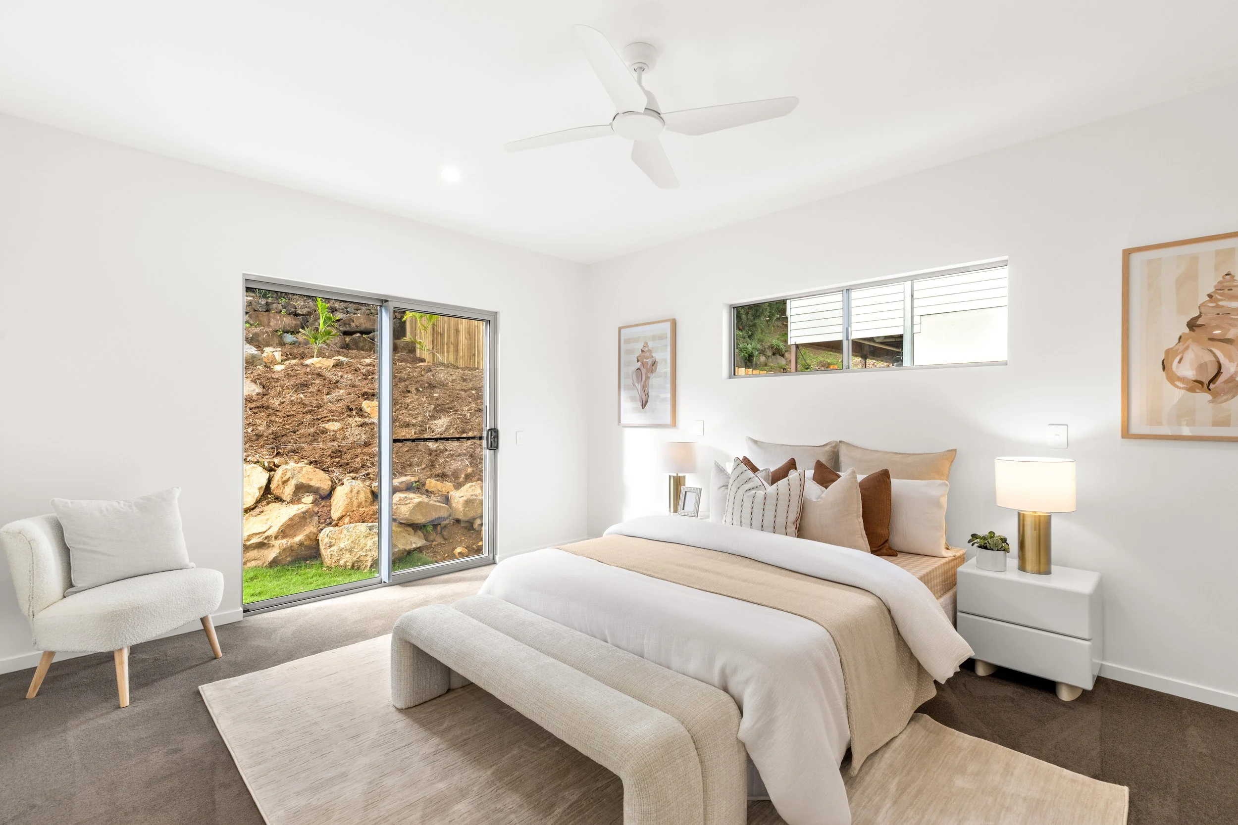 Bright living room with white walls, large windows, modern artwork, beige sofa, white armchair, and sliding glass door overlooking a garden with rocks and plants.