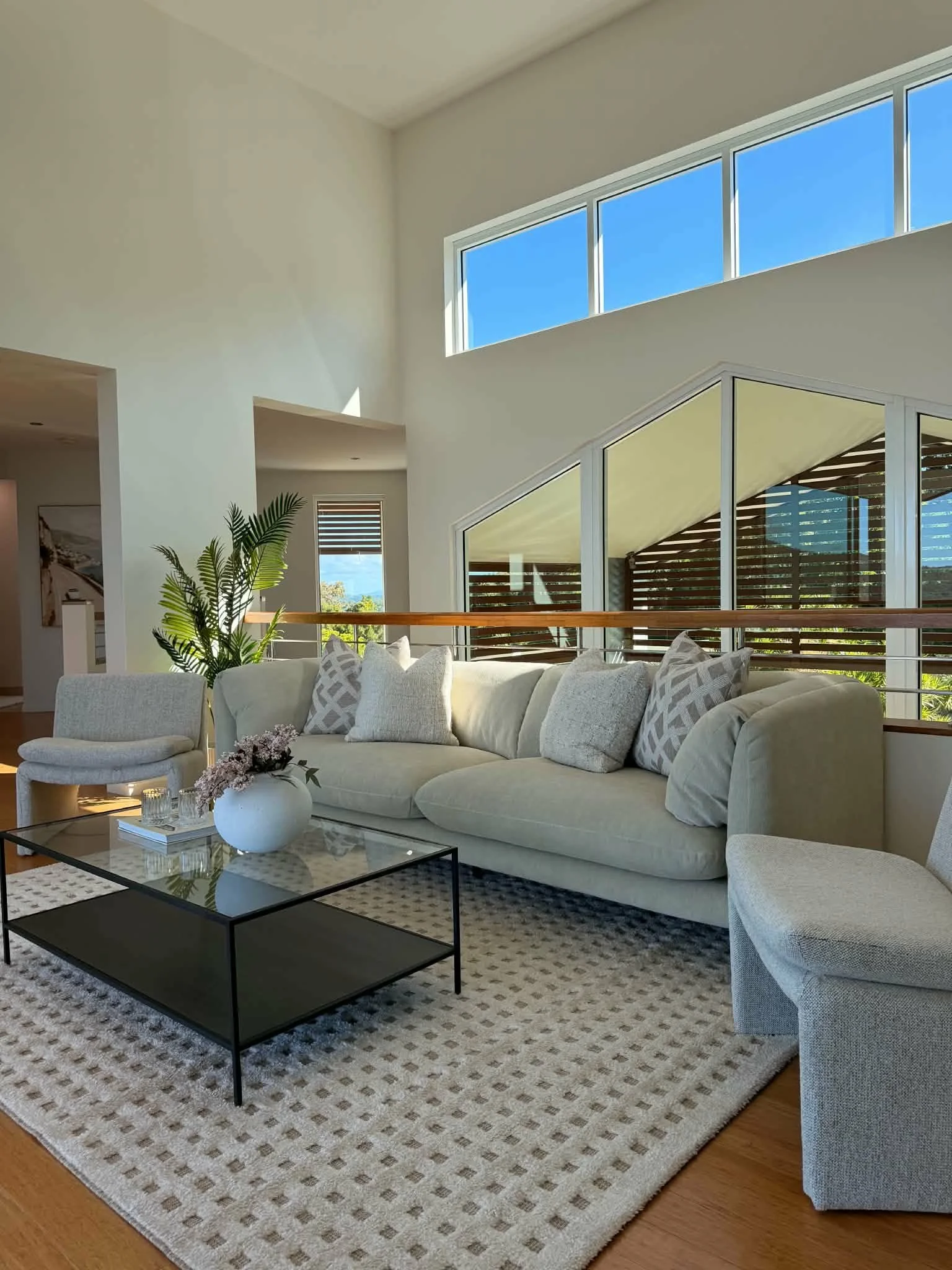 Bright living room with cream-colored sectional sofa, patterned throw pillows, a black coffee table, and a textured area rug. Large windows with views of greenery and a wooden balcony are visible, along with a potted plant and a framed piece of art on the wall.