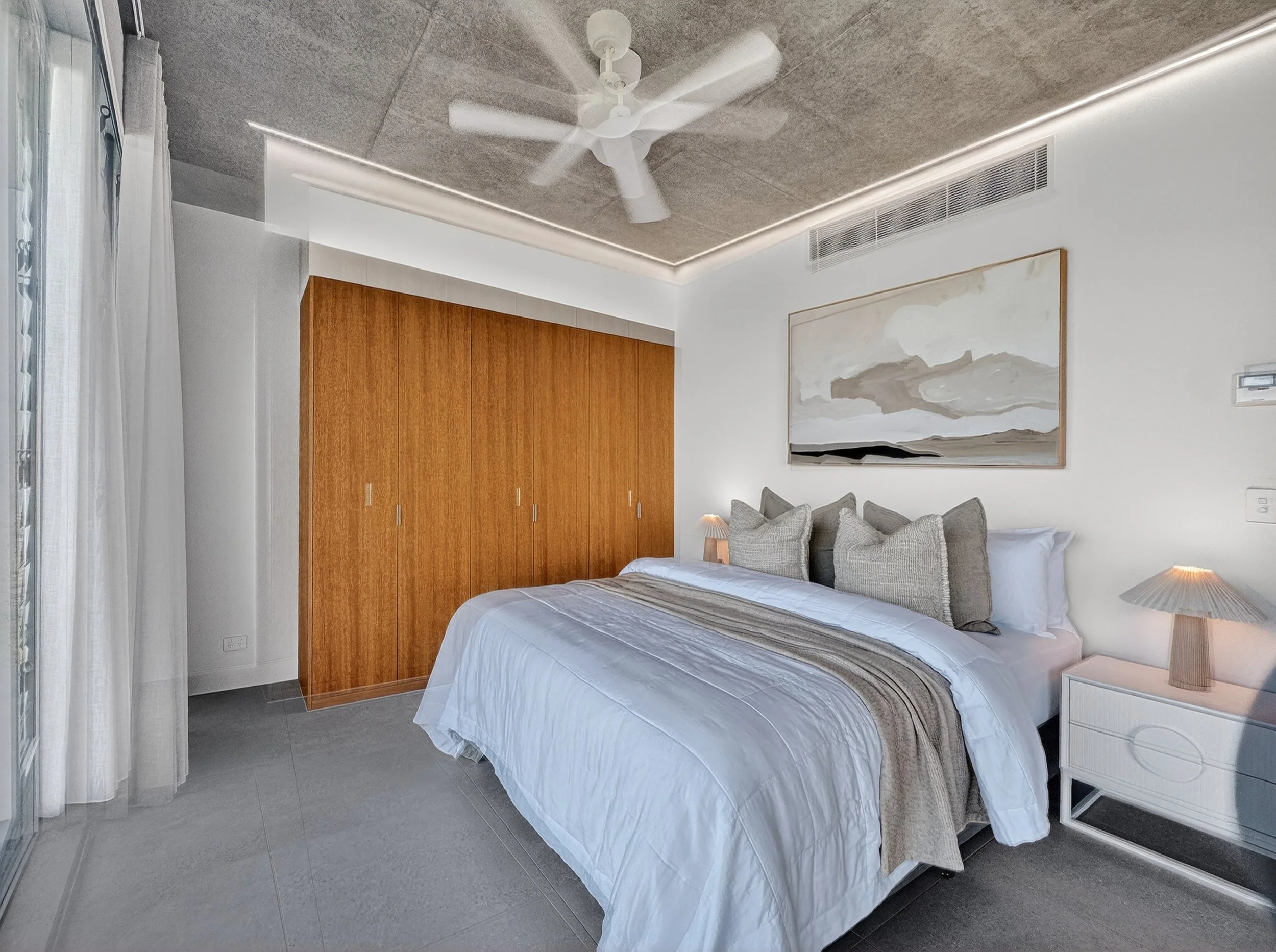 A bedroom with a white bed, gray pillows, and a bedside table with a lamp. There is a large wooden wardrobe, a piece of abstract artwork on the wall, a ceiling fan, and LED strip lighting on the ceiling.