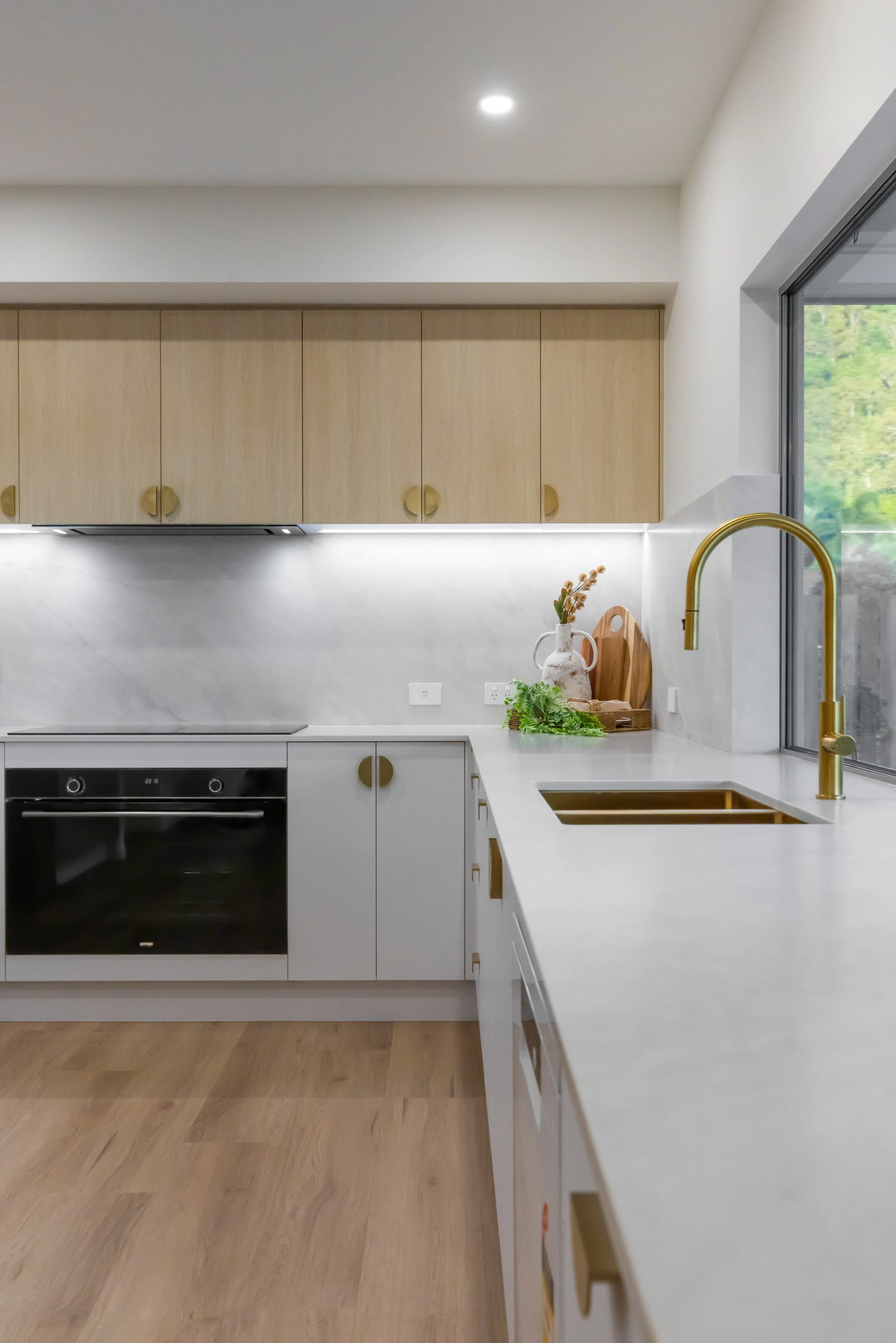 Modern kitchen with white countertops, light wood cabinets, a black oven, and a large window with a view of outdoor greenery.