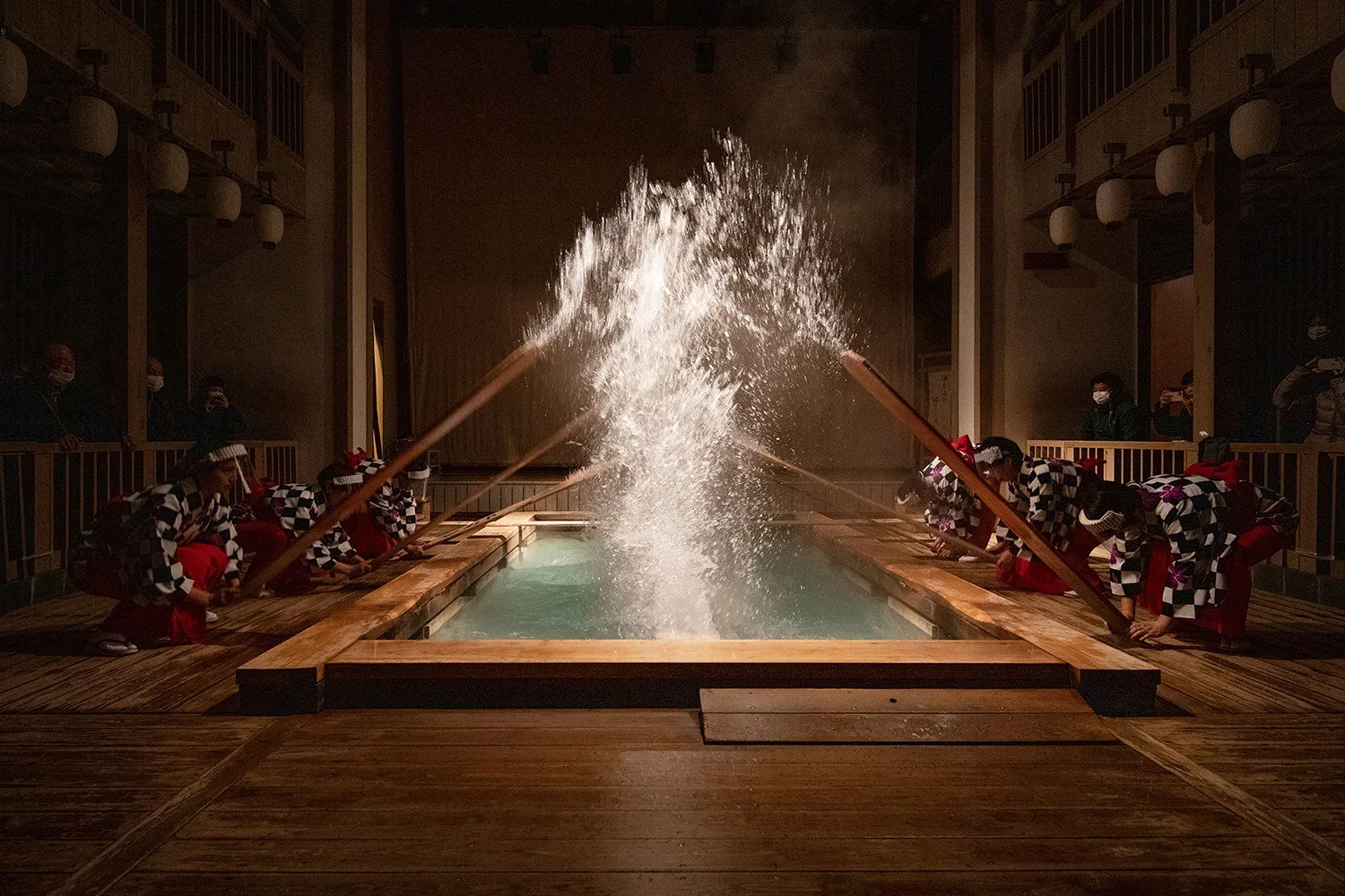 People dressed in traditional Japanese clothing kneeling around a wooden hot spring bath, splashing water from long wooden poles during a ceremony at night.