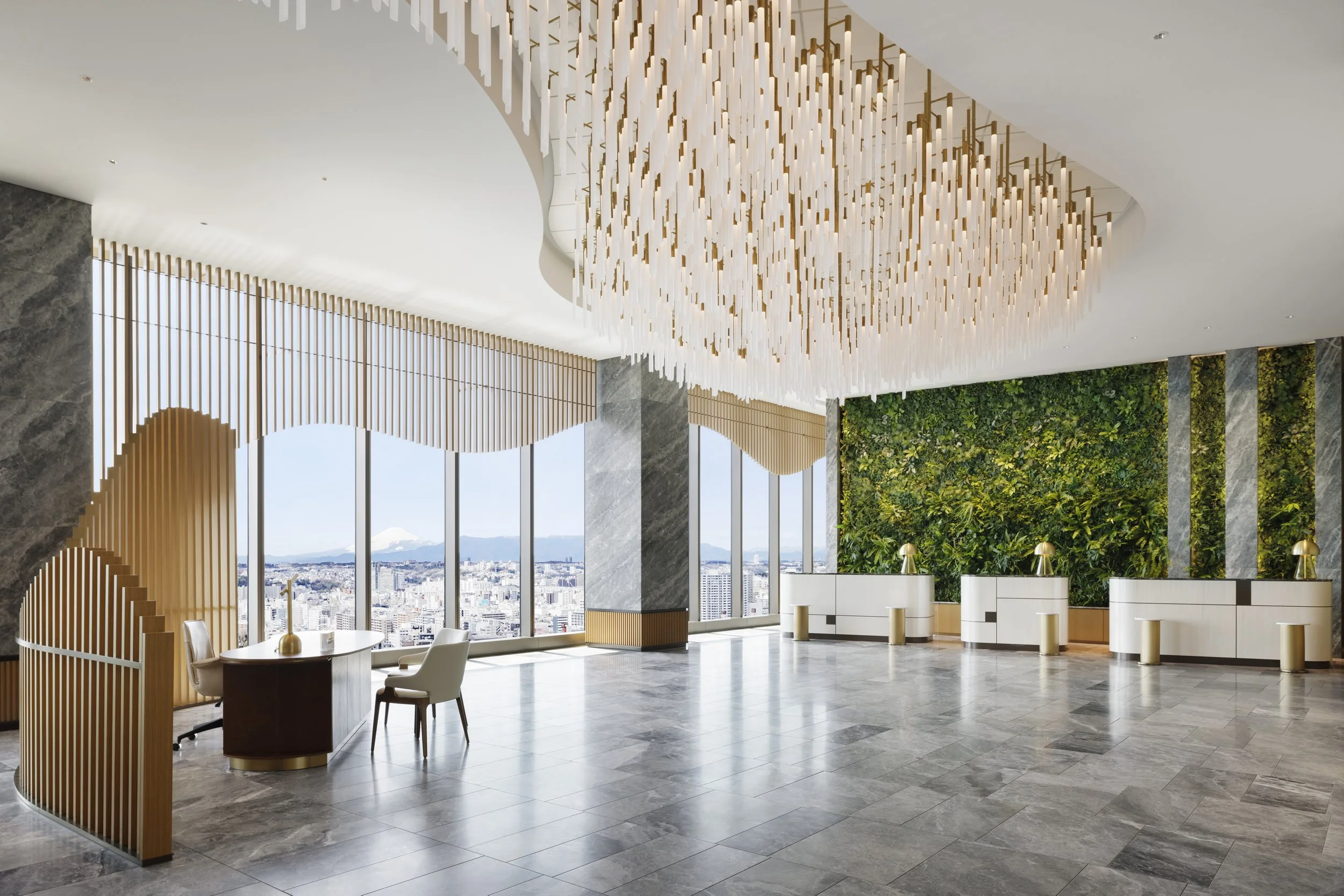 Modern office lobby with large windows, a green wall, a wood and white reception desk, and a contemporary chandelier made of dangling white and gold elements.