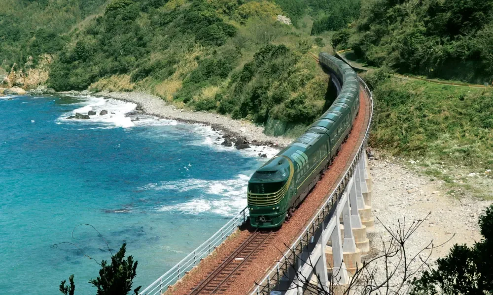A green train traveling along a track on a bridge over ocean water, with green hills and rocky shoreline.