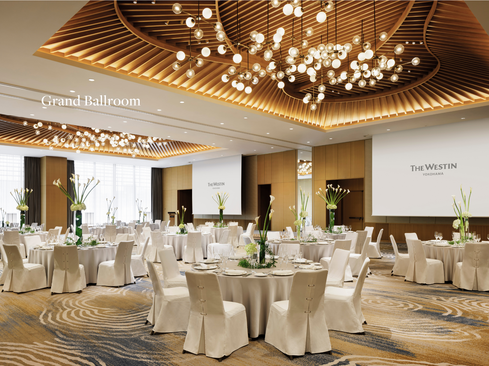 A luxurious hotel ballroom decorated for an event, with round tables covered with white tablecloths, elegant chairs, tall floral centerpieces with calla lilies, and large screens displaying 'The Westin Yokohama' branding. The ceiling features intricate wooden paneling and modern chandeliers.
