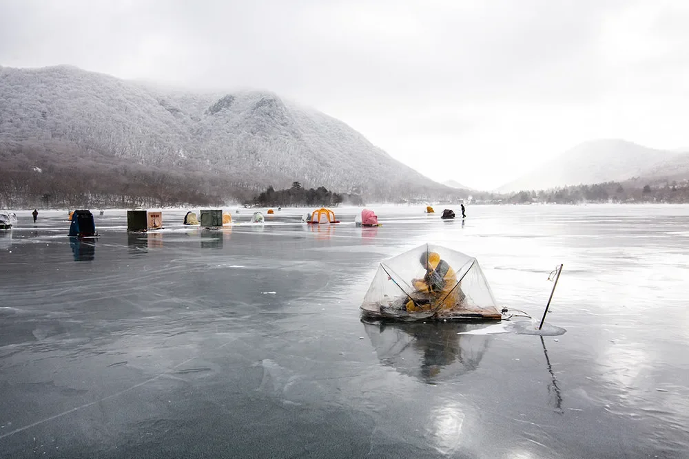Person ice fishing on a frozen lake with mountains in the background, surrounded by several other ice fishing huts.