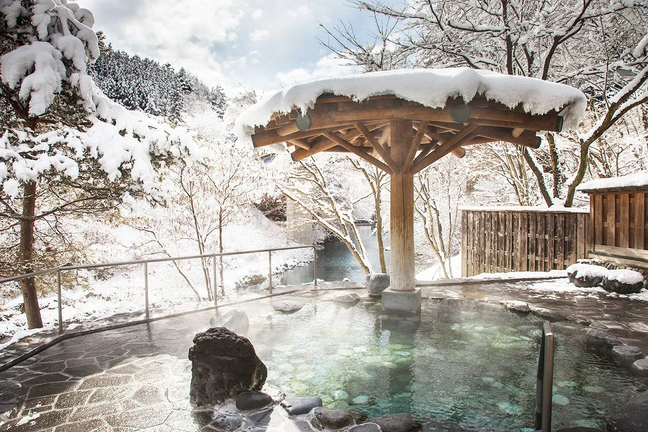 Steaming outdoor hot spring bath with wooden umbrella structure during winter, snow-covered trees and landscape in the background, and a fenced wooden area.
