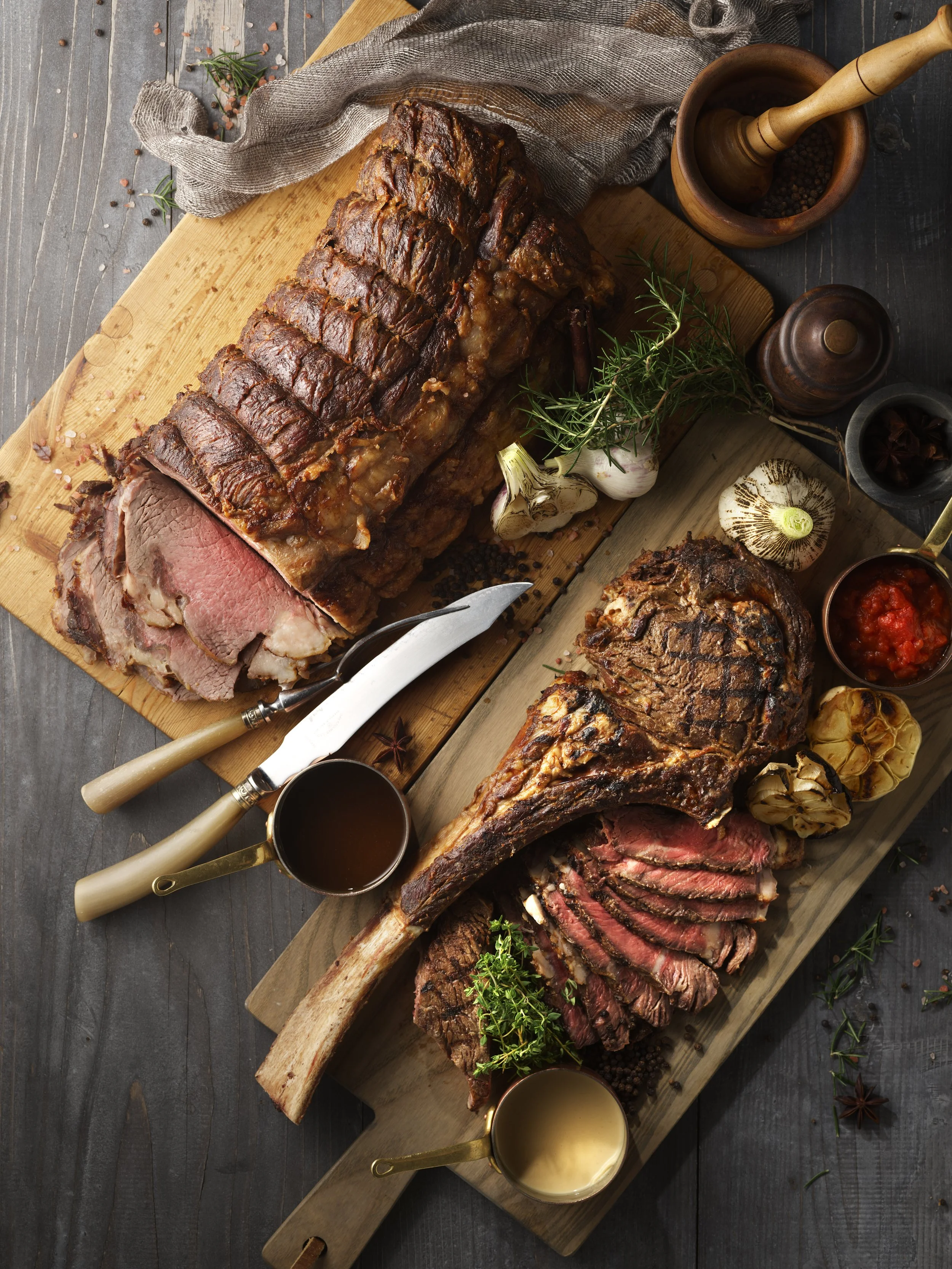 Two large cuts of cooked ribs on wooden cutting boards, with sliced meat exposed, garnished with herbs, surrounded by garlic, spices, and sauce.