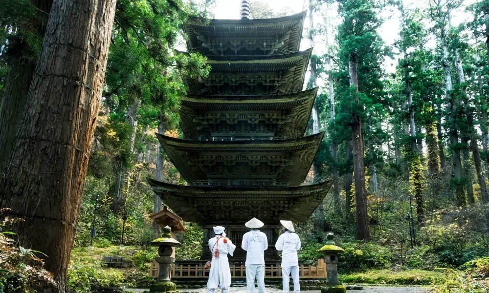 Three people dressed in traditional white attire stand facing a five-tiered pagoda in a forested area with tall trees.