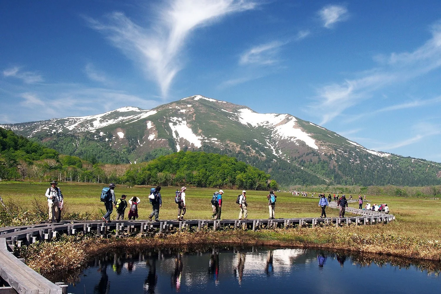 Group of hikers walking along a curved wooden path in a scenic landscape with snow-capped mountains, green trees, and a pond reflecting the sky.