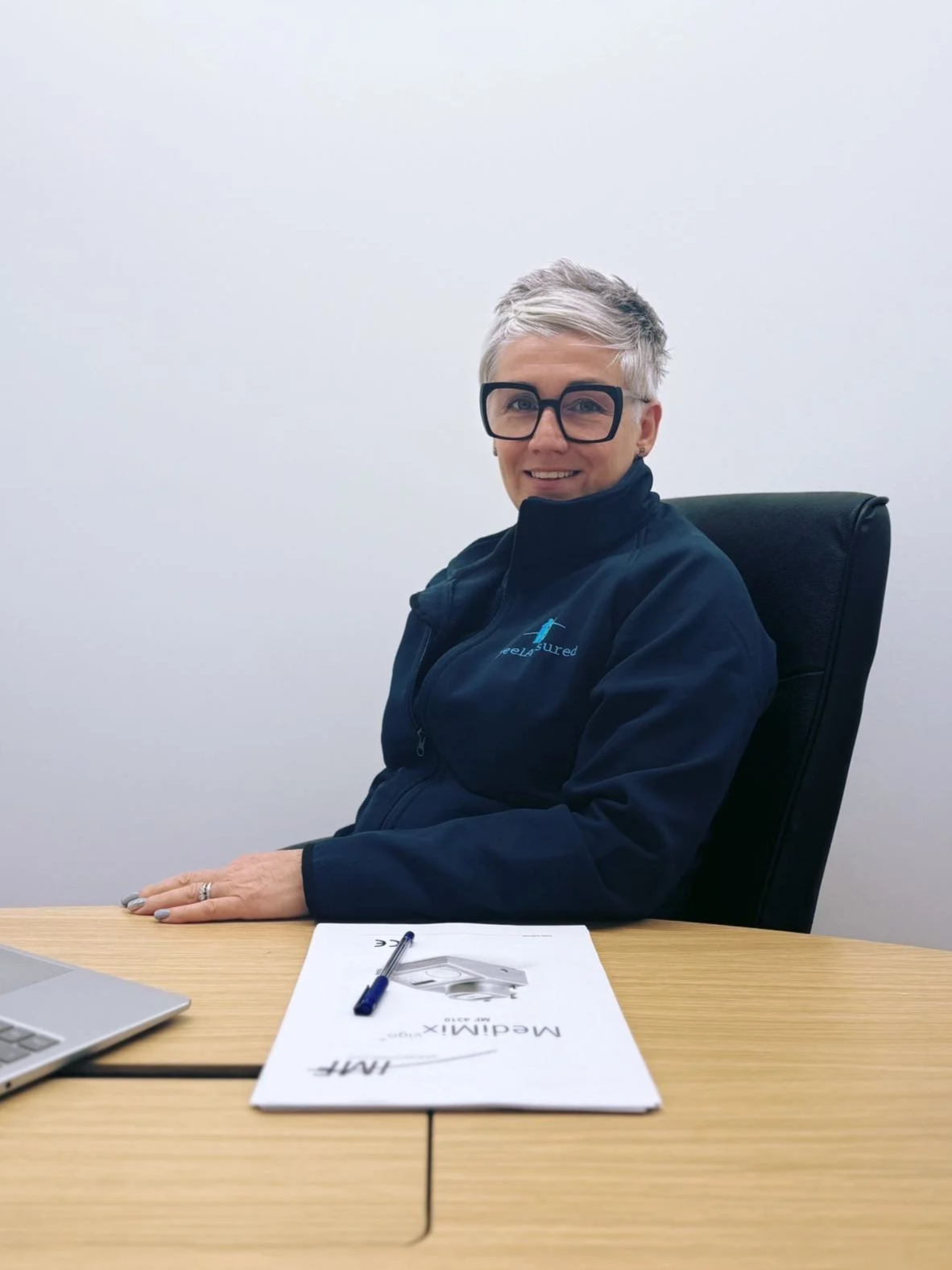 A woman with short gray hair, wearing large black glasses and a dark jacket, is sitting at a wooden desk in an office. There are some papers, a pen, and a laptop on the desk. The woman is smiling and looking at the camera.