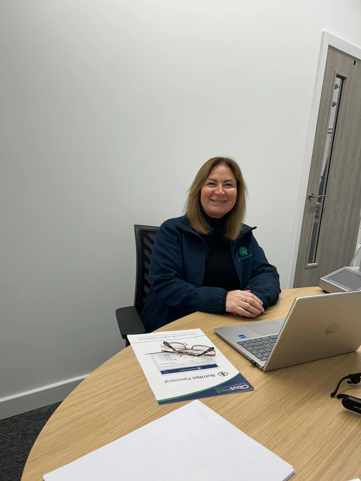A woman with shoulder-length light brown hair sitting at a round wooden desk, smiling at the camera, with eyeglasses, a laptop, pamphlets, and papers in front of her, in an office with white walls and a door in the background.