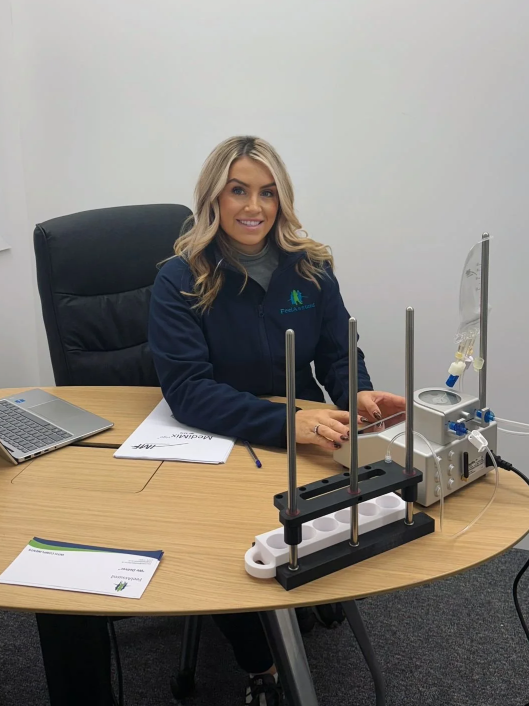 A woman in a navy jacket sitting at a desk with lab equipment and a laptop, smiling at the camera.