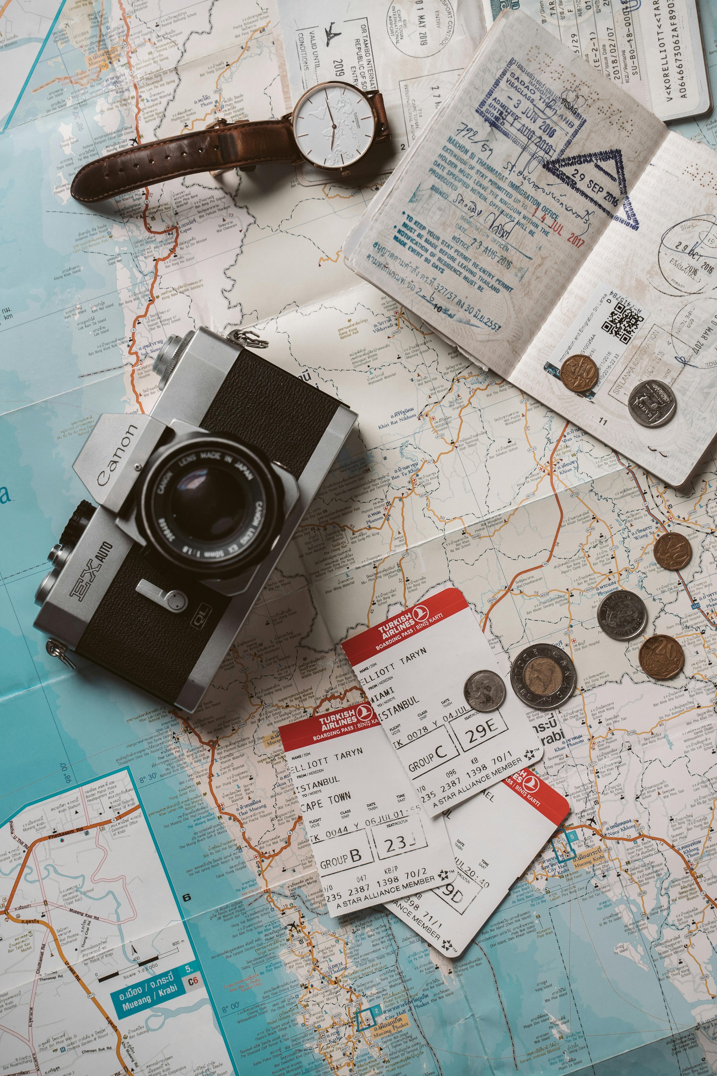 Travel essentials on a map, including a vintage camera, a wristwatch, travel documents like tickets and passport, coins, and a boarding pass.