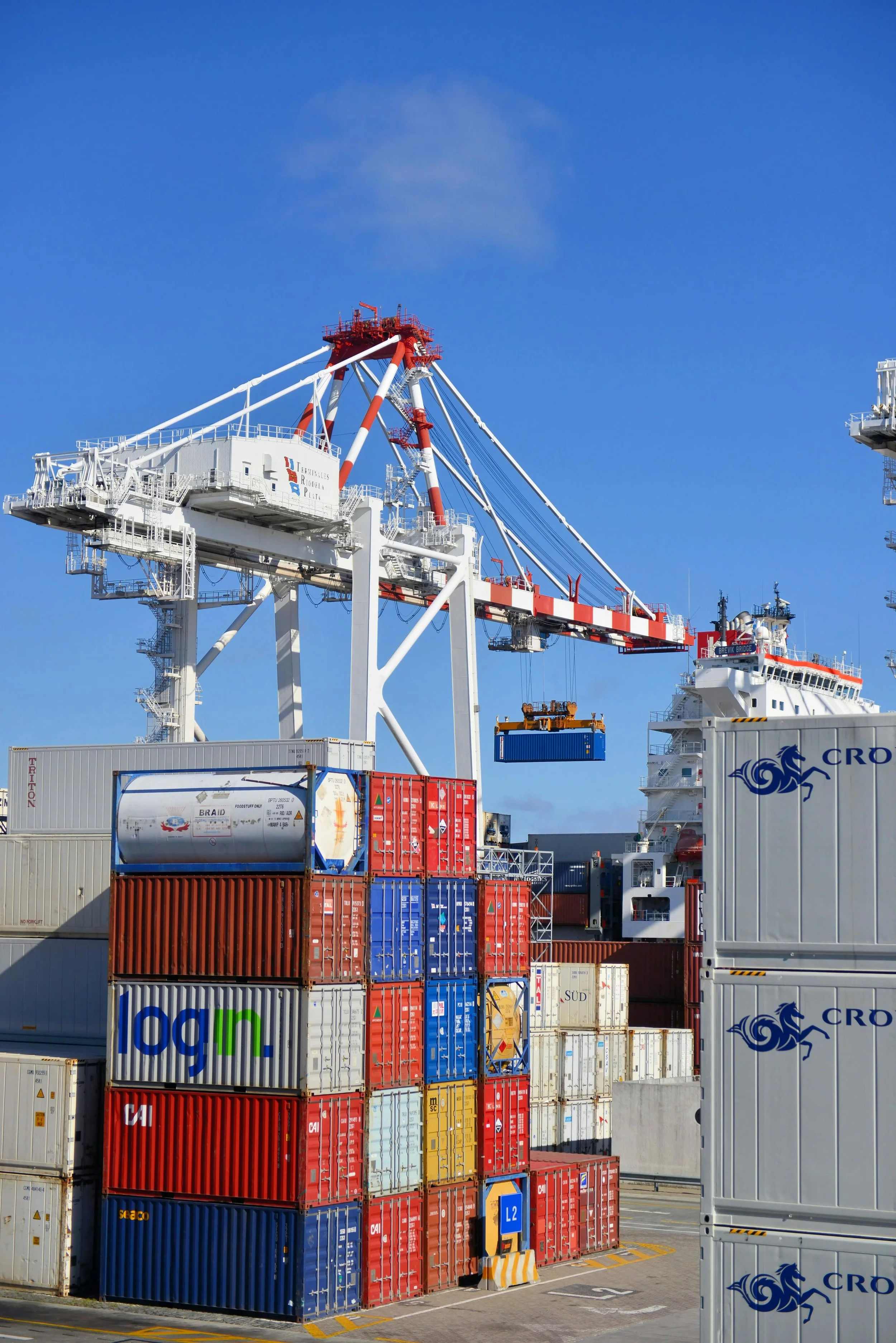 Shipping containers stacked at a port with a large crane overhead, ships visible in the background.
