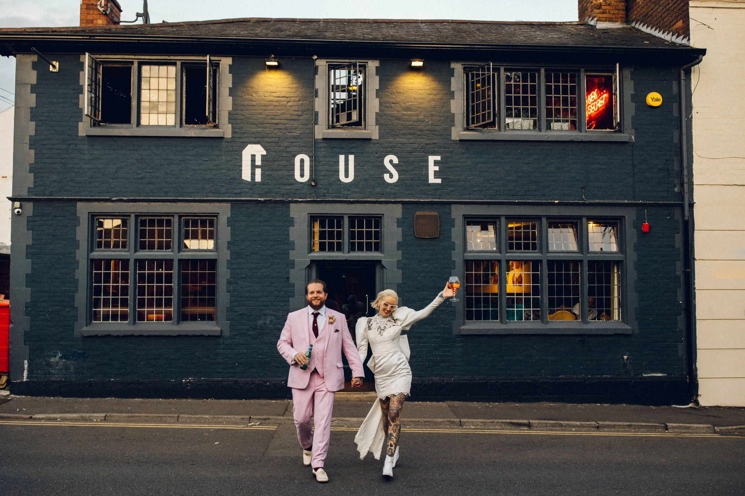 A groom in a pink suit holding a beer holds hands with his bride in a short mini wedding dress, tattoos and holding an Aperol spritz dancing in front if their pub wedding venue.