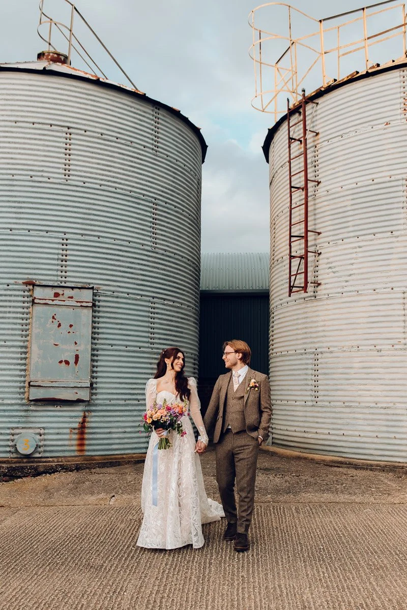 A bride and groom walks holding hands in front of two metal silos, the bride holds a bouquet of spring flowers with blue ribbon