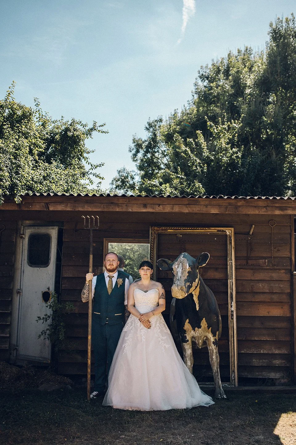 A man and woman in wedding attire standing outdoors in front of a wooden shed with a cow sculpture and an empty picture frame behind them, under a bright blue sky with trees.