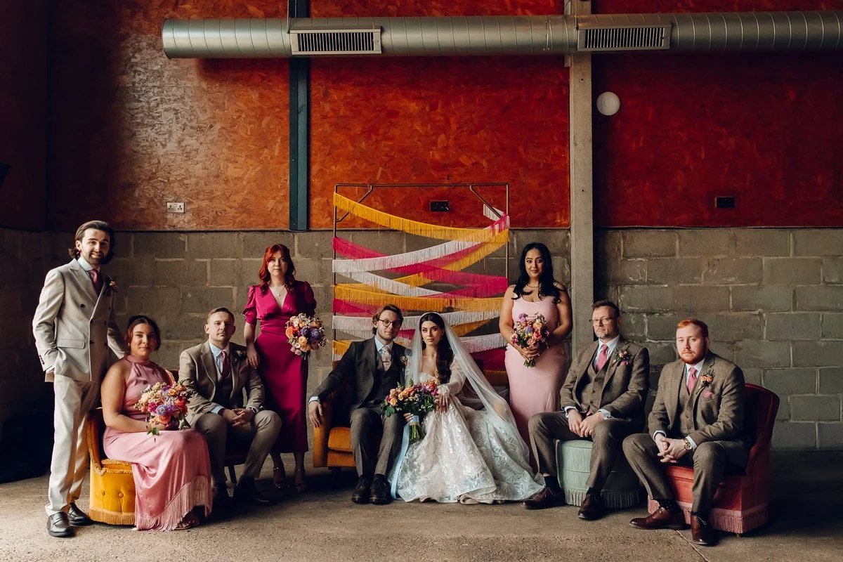 A moody group portrait of he bride and groom and wedding party sit and stand on sofas infront of a vintage tassels backdrop