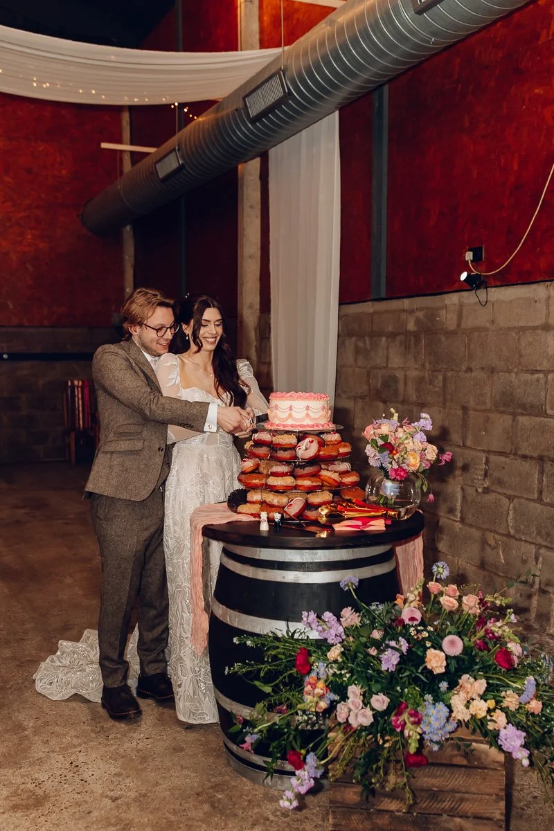  A groom puts his arm around the bridge whilst they cut a cake that sits on a tower of donuts on a barrel.  