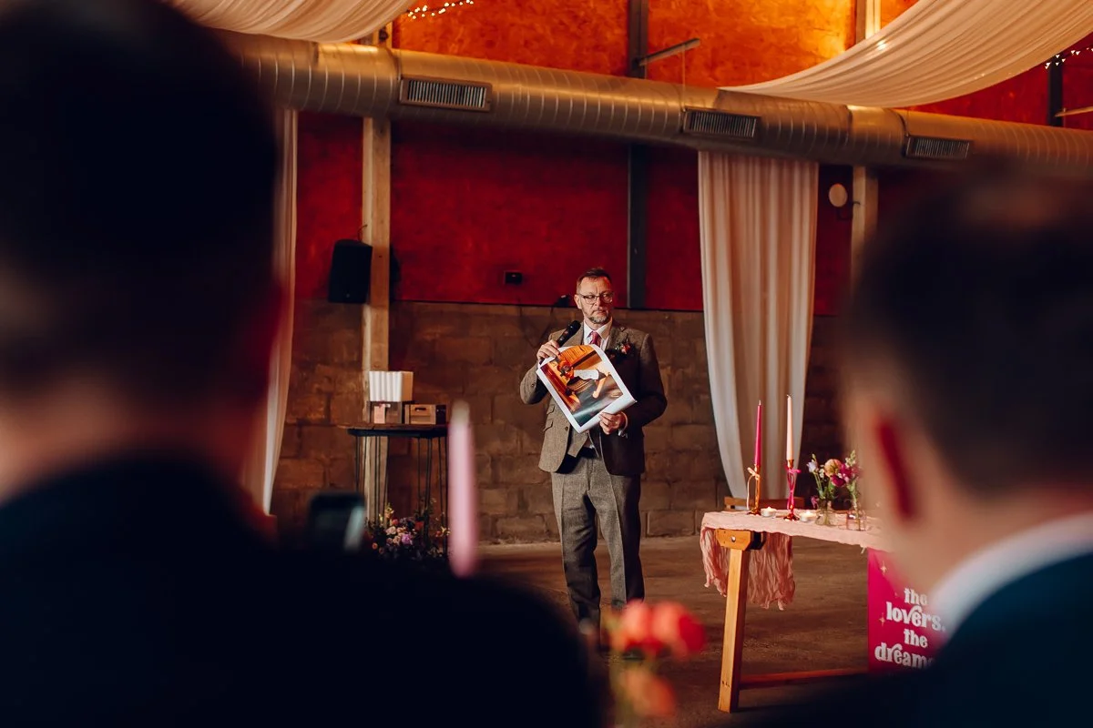  A man holds up a photo of the groom during his speech at lodge farm barns.  