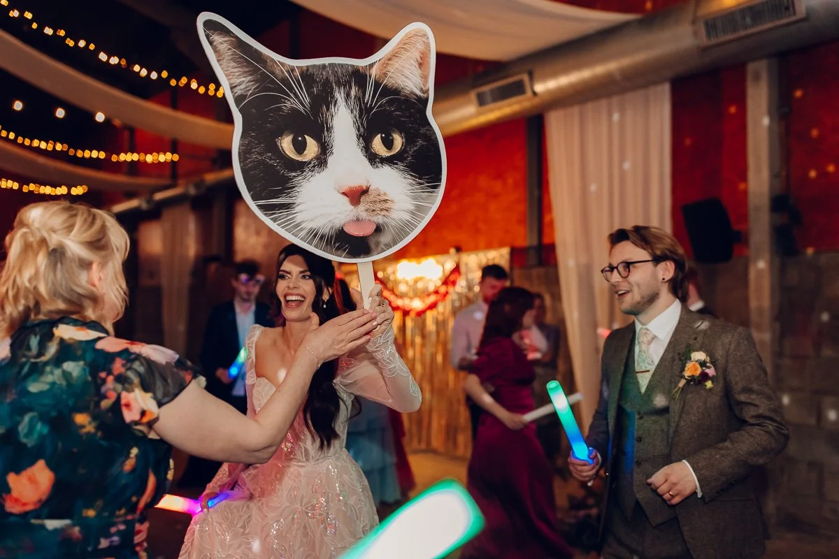  The bride and groom laugh on the dance floor whilst the bride holds up a giant cut out of their cats face 