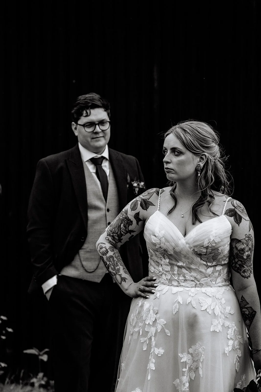 A black and white image of a couple standing in front a black barn door. The groom stands behind the bride looking at her whilst the bride wearing a floral dress with tattooed arms looks to the left