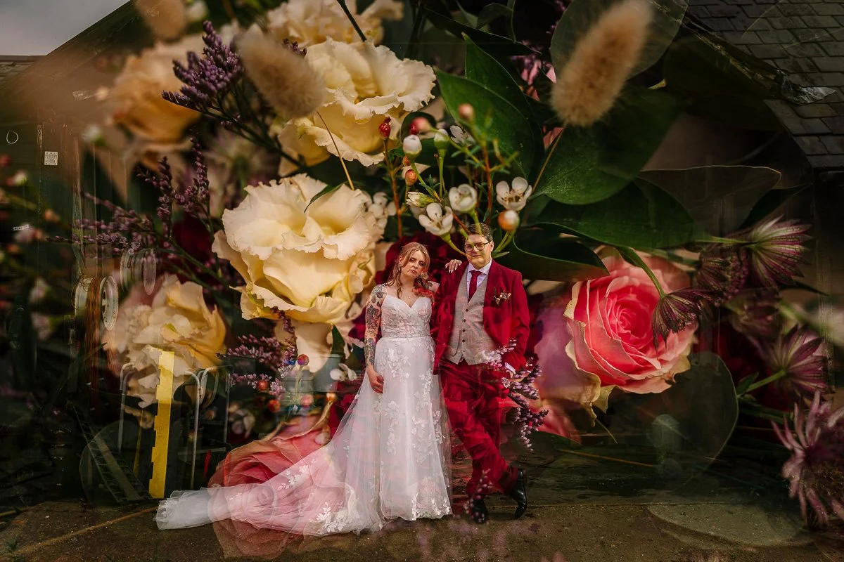 A bride in a white dress with tattoos and a groom with a red suit stand staring at the camera whilst an Image of flowers has been exposed on top