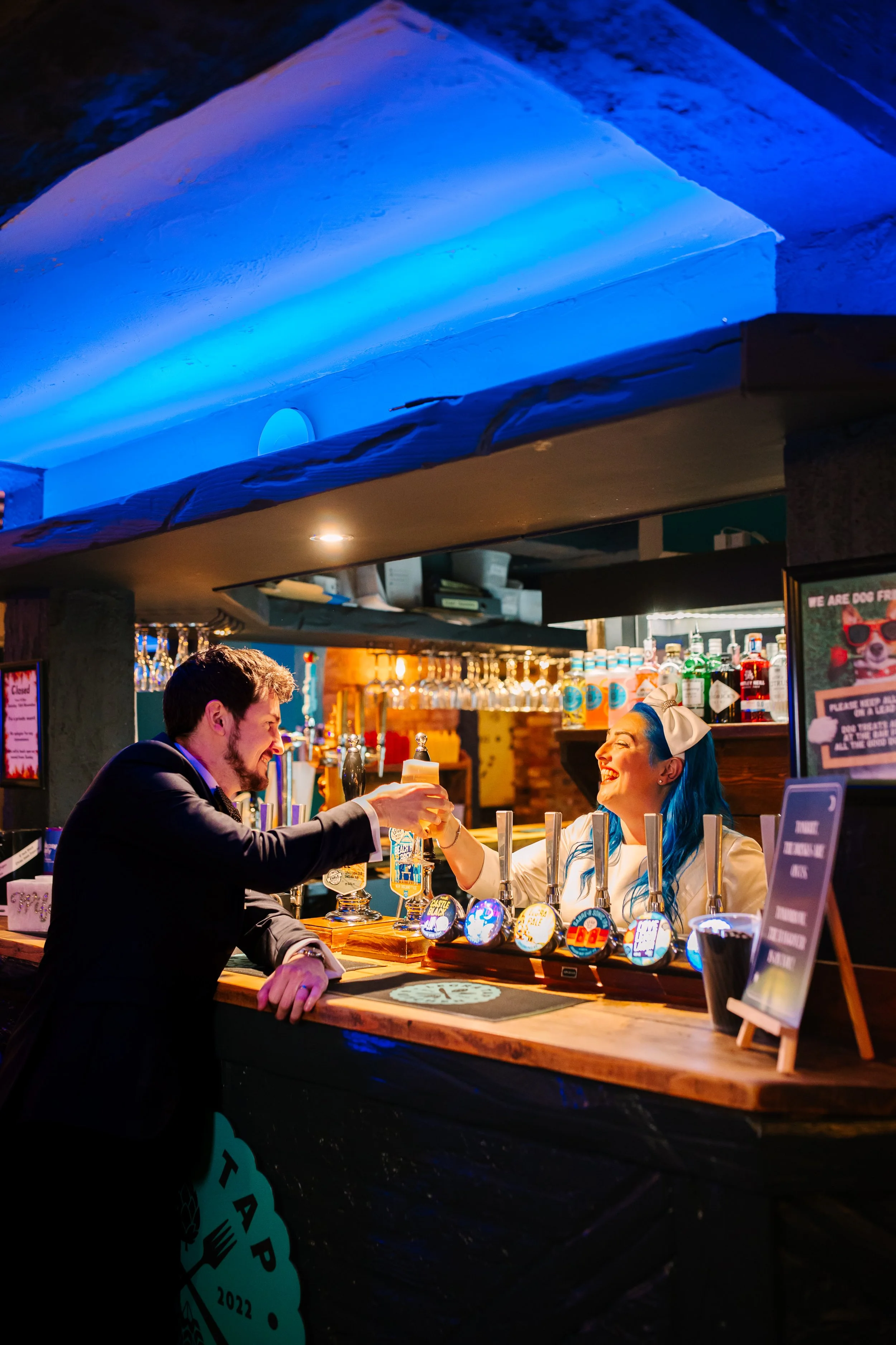 A bride with blue hair stands behind the bar in a pub and offers her husband a pint of beer.