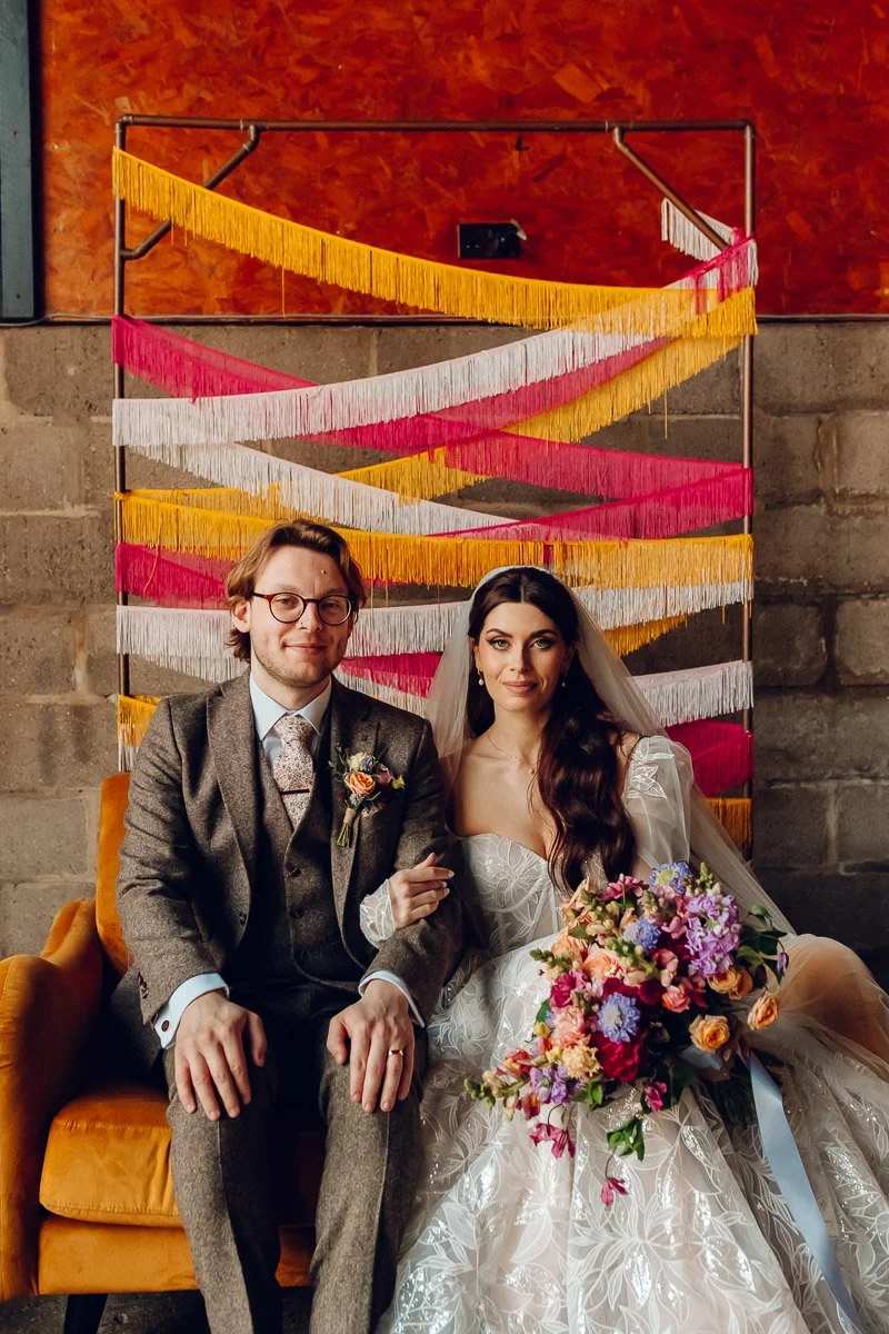 a groom in a brown tweed suit sits next to his bride on a mustard sofa infront of a vintage style yellow and pink fringed backdrop