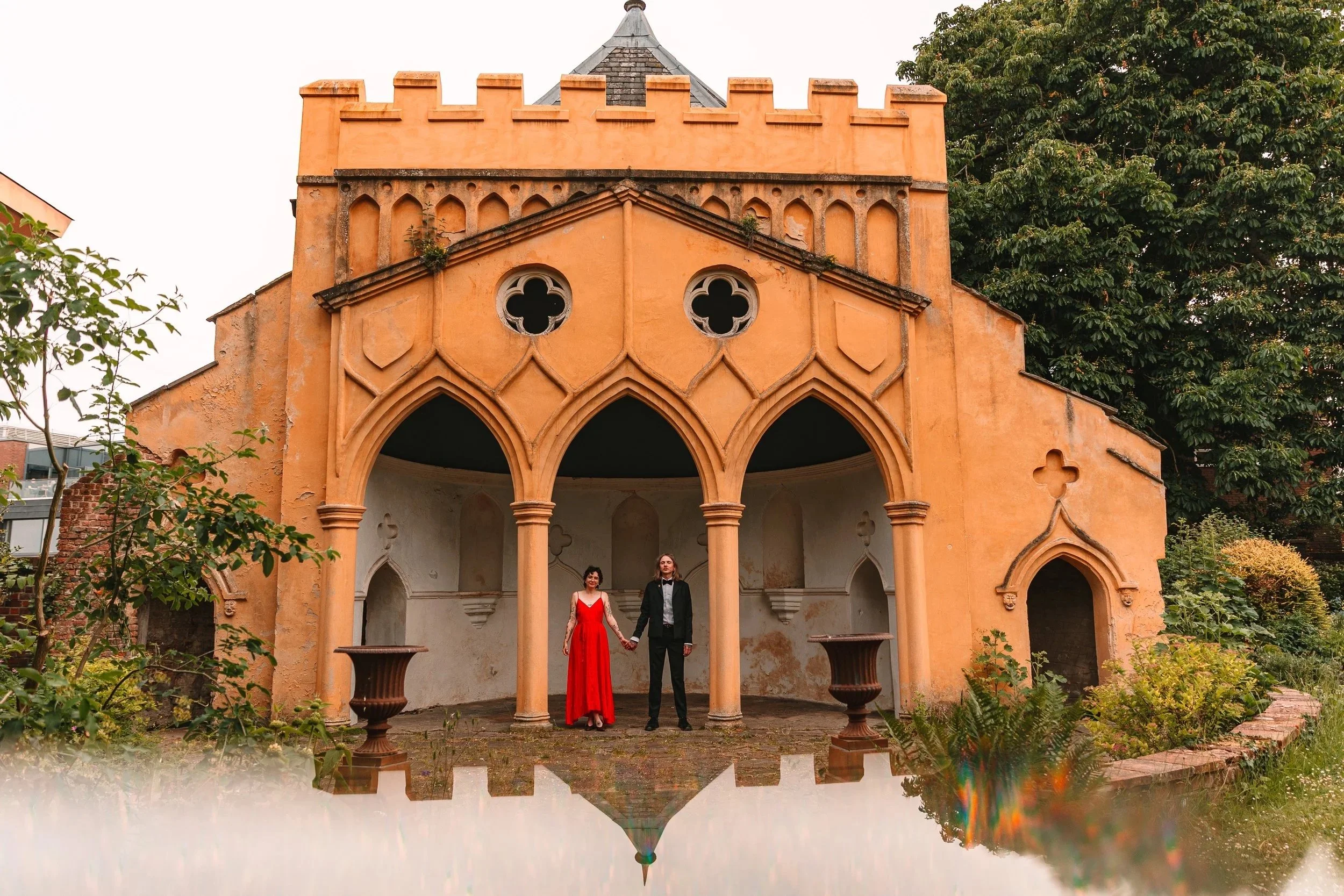 A bride in a red dress holds hands with her husband wearing a black tuxedo underneath a gothic folly at the minors colchester