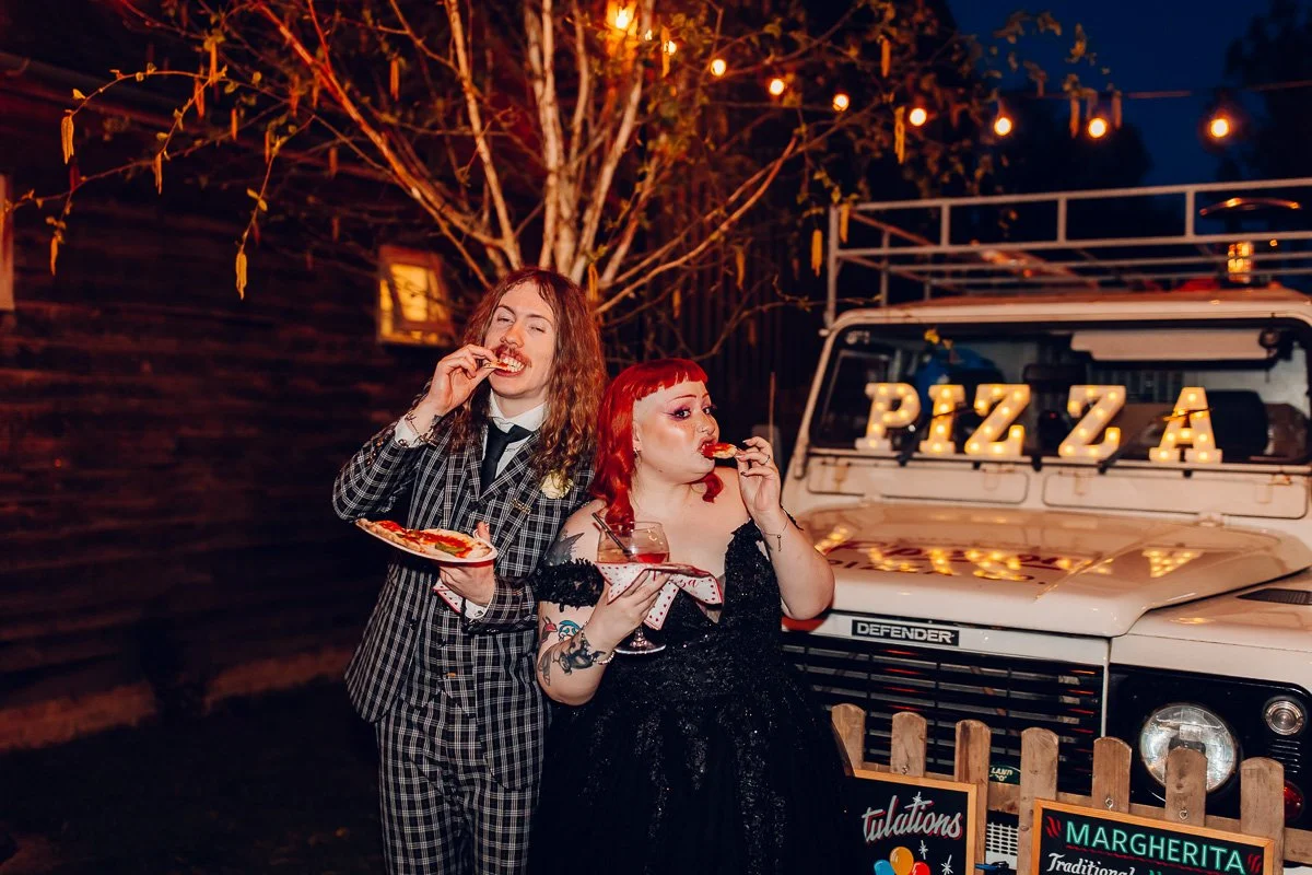 A groom in a black checked suit and bride in a black dress stand infront of a pizza van eating pizza