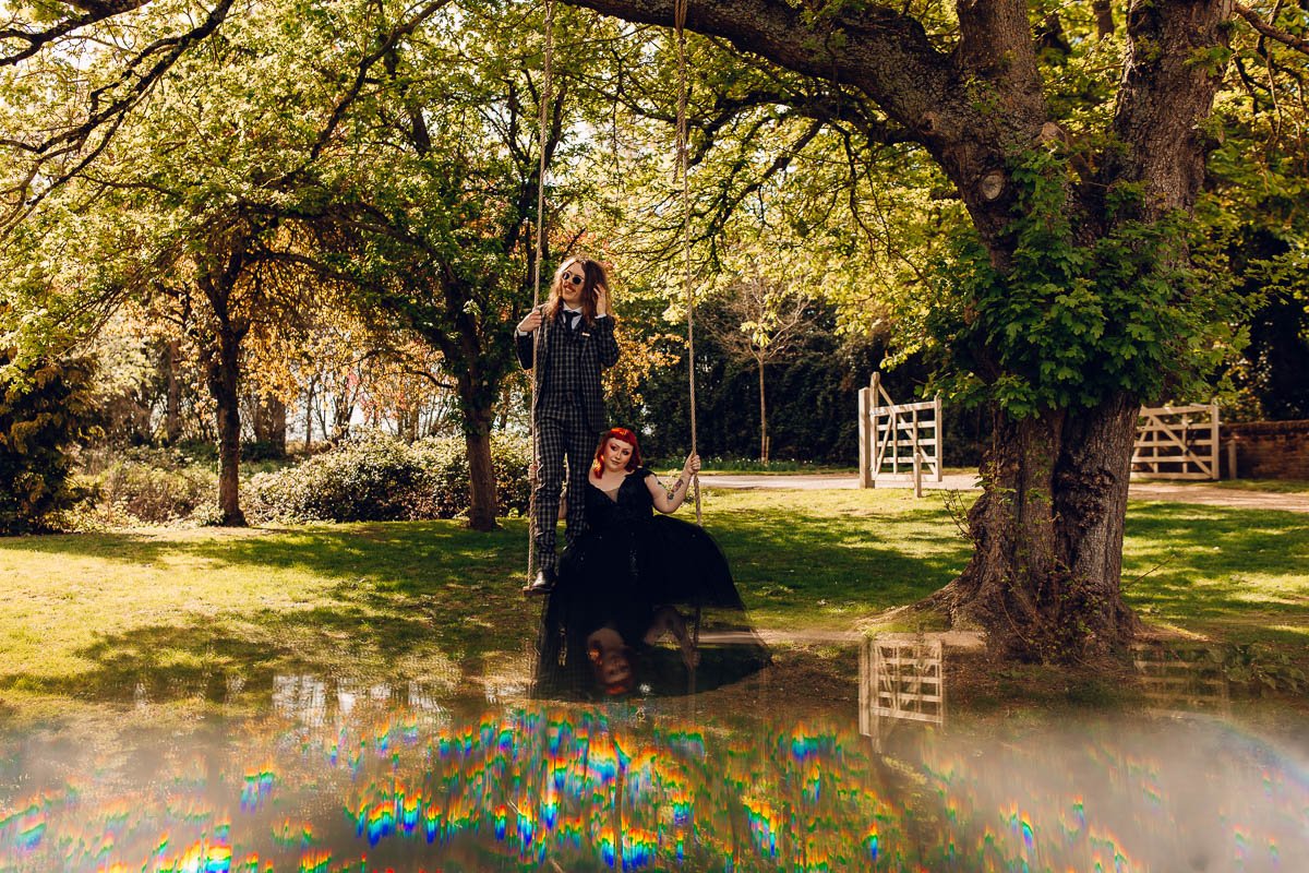 A bride with red hair wears a black wedding dress whilst sitting on a swing, the Groom stands on the swing next to her in a black checked suit