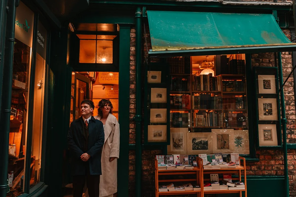 A groom in a navy coat and bride in a beige coat stand in a bookshop door way on the York elopement