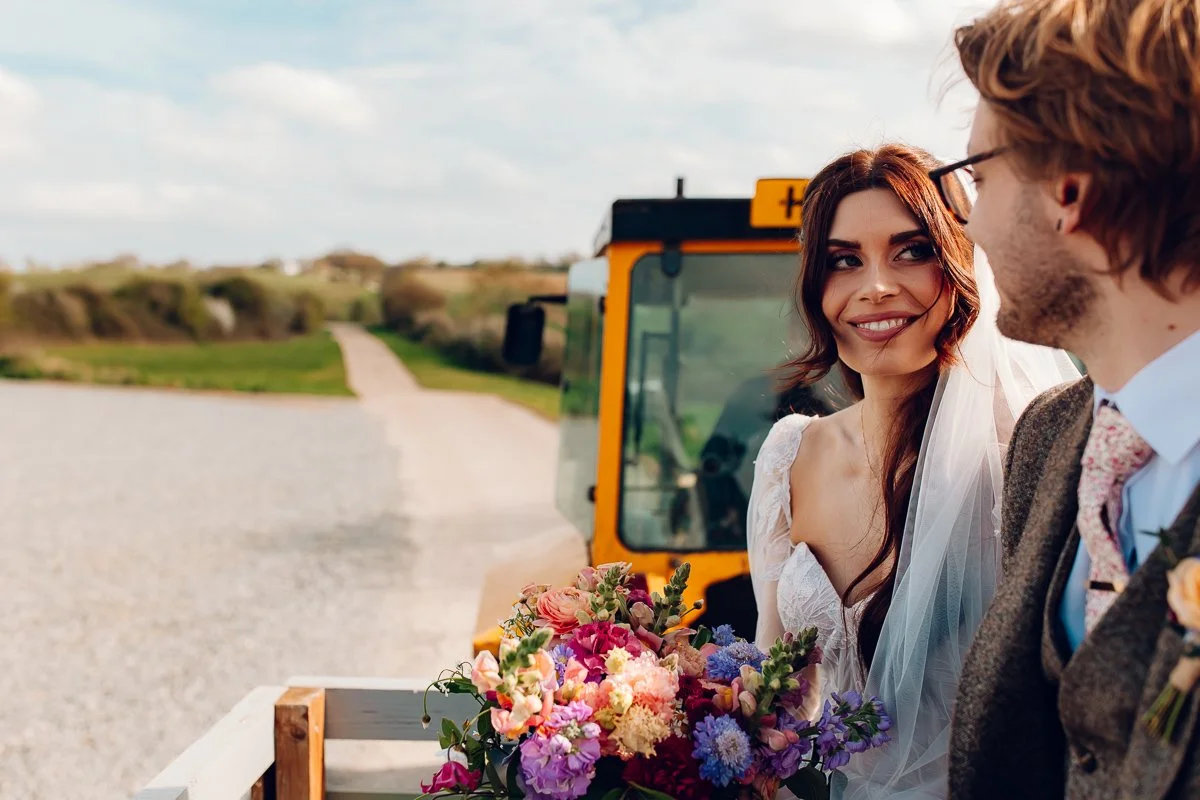 Bride and groom sit on the back of a trailer attached to a yellow tractor, the bride smiles at the groom