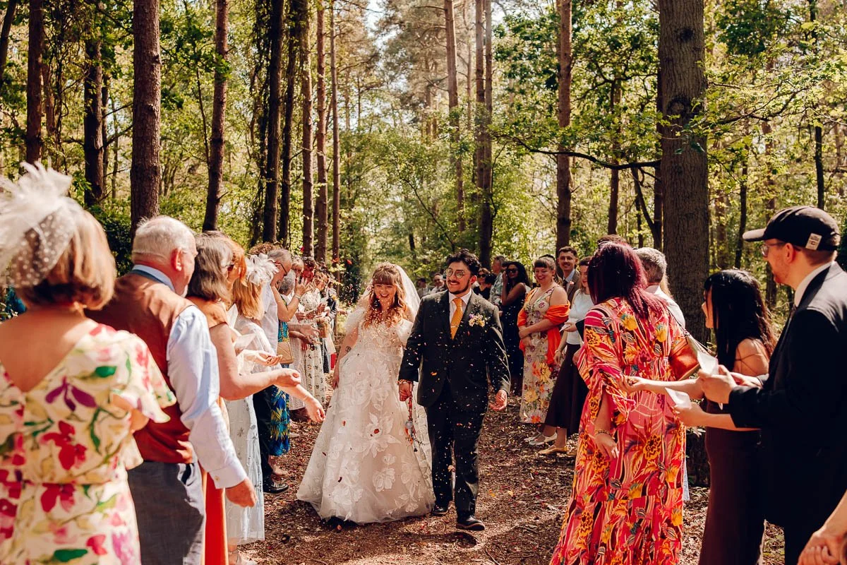 Amongst a woodland path lined with trees a bride and groom walk hang in hand whilst guests through confetti over them at captains wood barn
