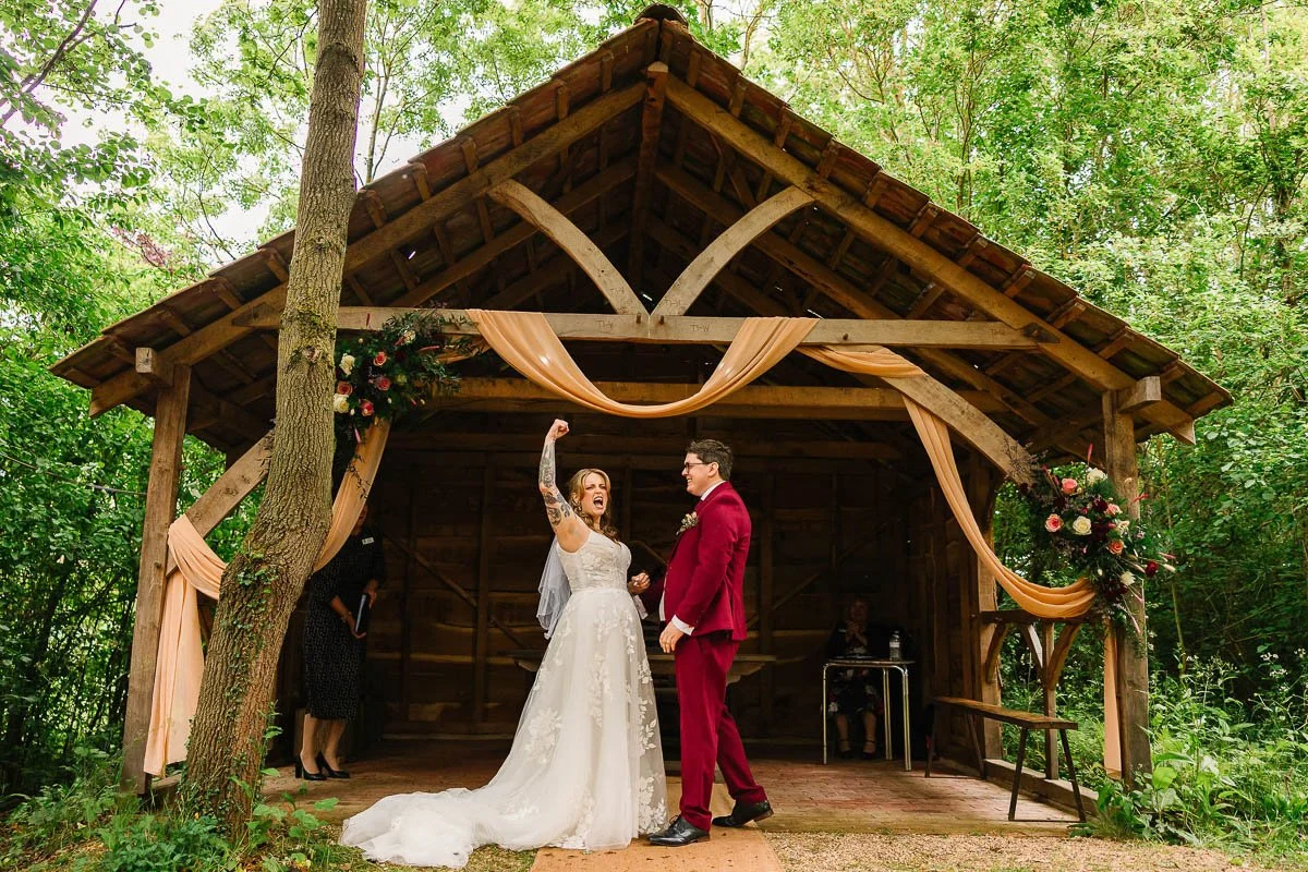A bride with tattoos in a white dress fist bumps the air whilst her new husband in a red suit laughs with her