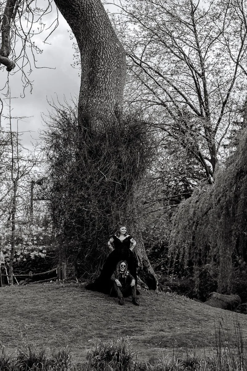 A black and white photo of a couple at Houchins, the bride stands with her hands on her hips in front of a tree whilst the groom sits on the floor