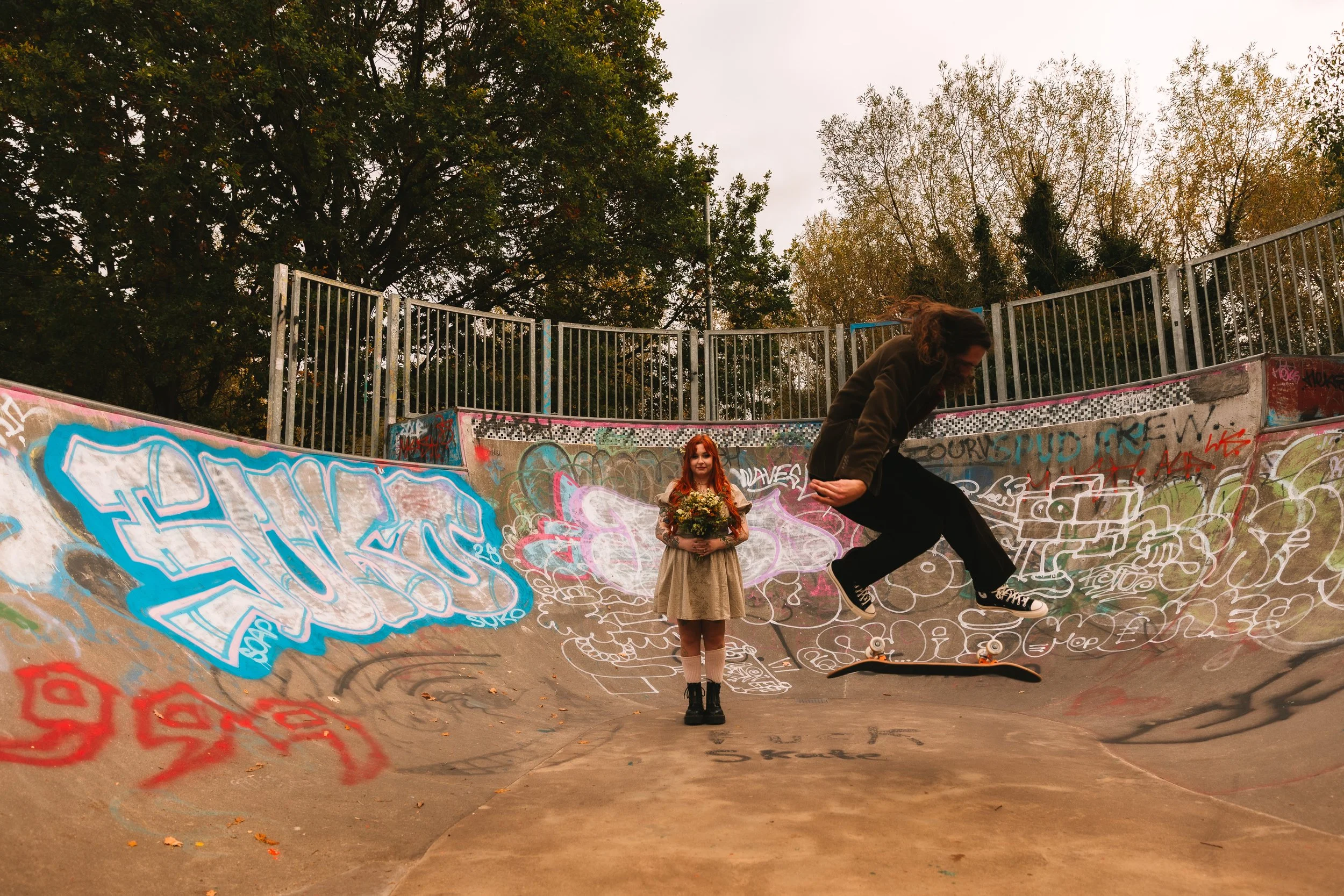 A bride with red hair in a short dress stands in a graffiti covered skatepark whilst her husband skates in front of her wearing converse