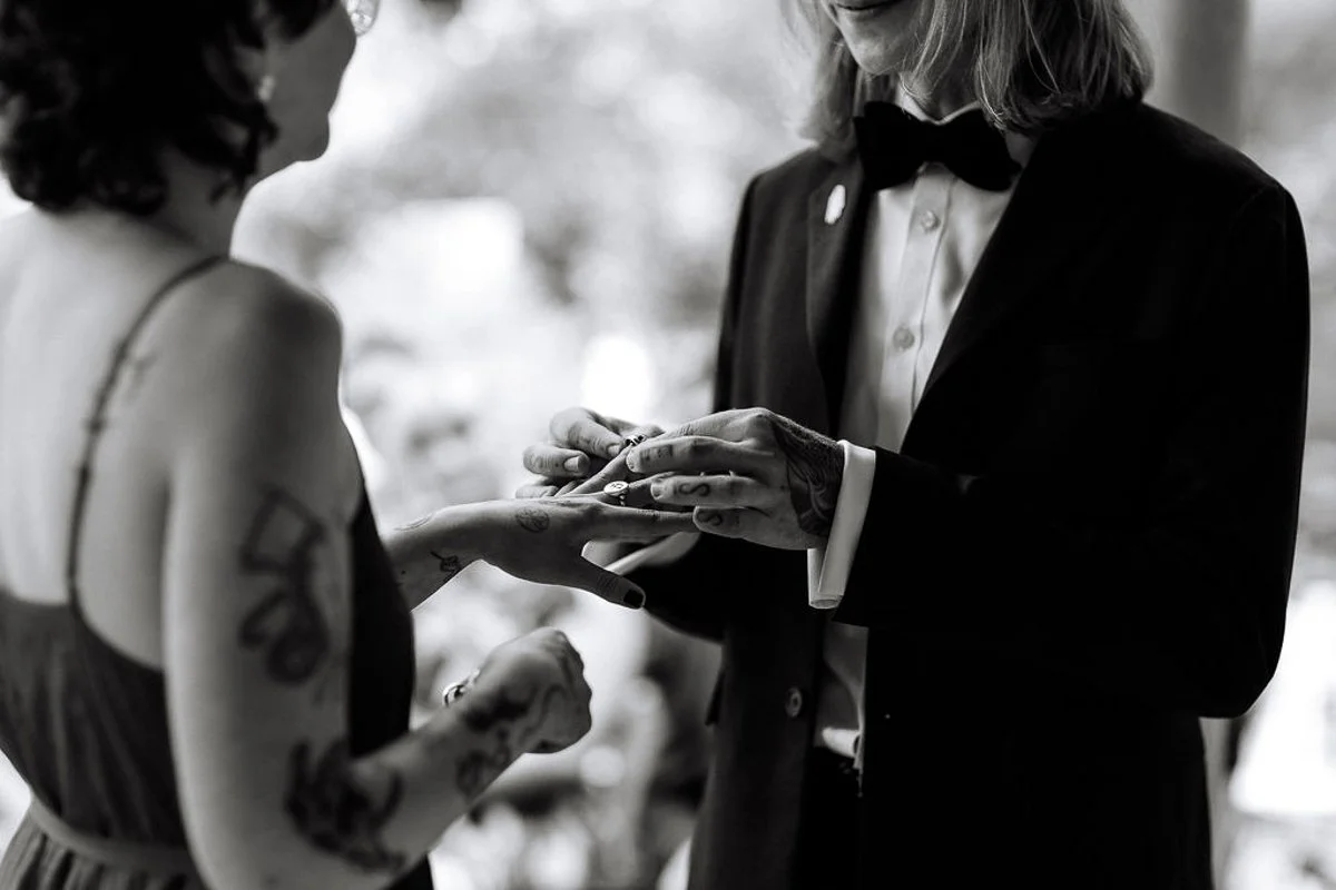 A Black and white photograph of a groom placing a wedding ring onto a bride with tattoos finger at the Minories