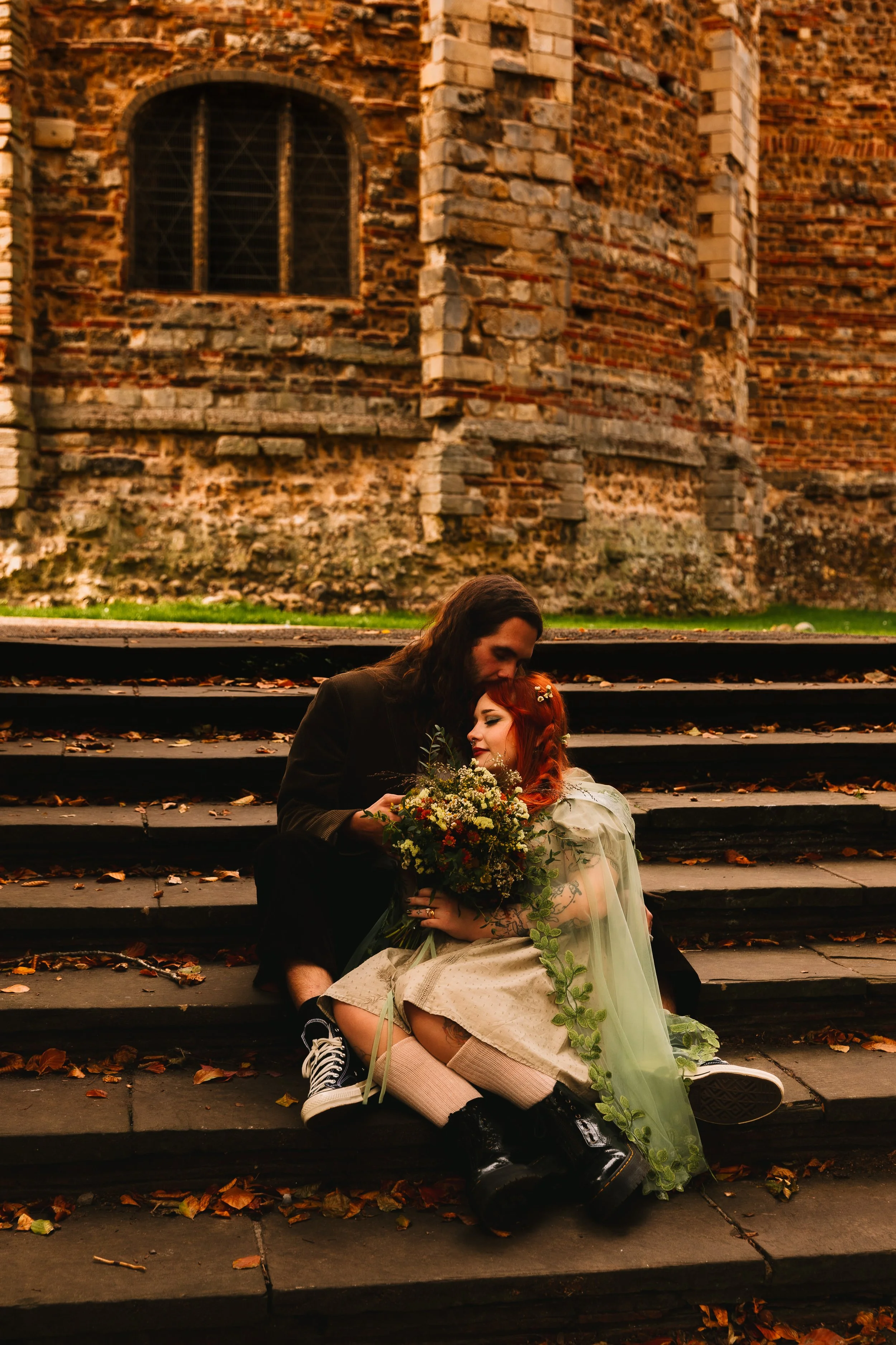 Alternative autumn wedding portrait of a couple sitting on stone steps at Colchester Castle Park in Essex. The bride has red hair, wears a green dress and cape and holds a wildflower bouquet. The Groom sits behind her wearing a suit and trainers.