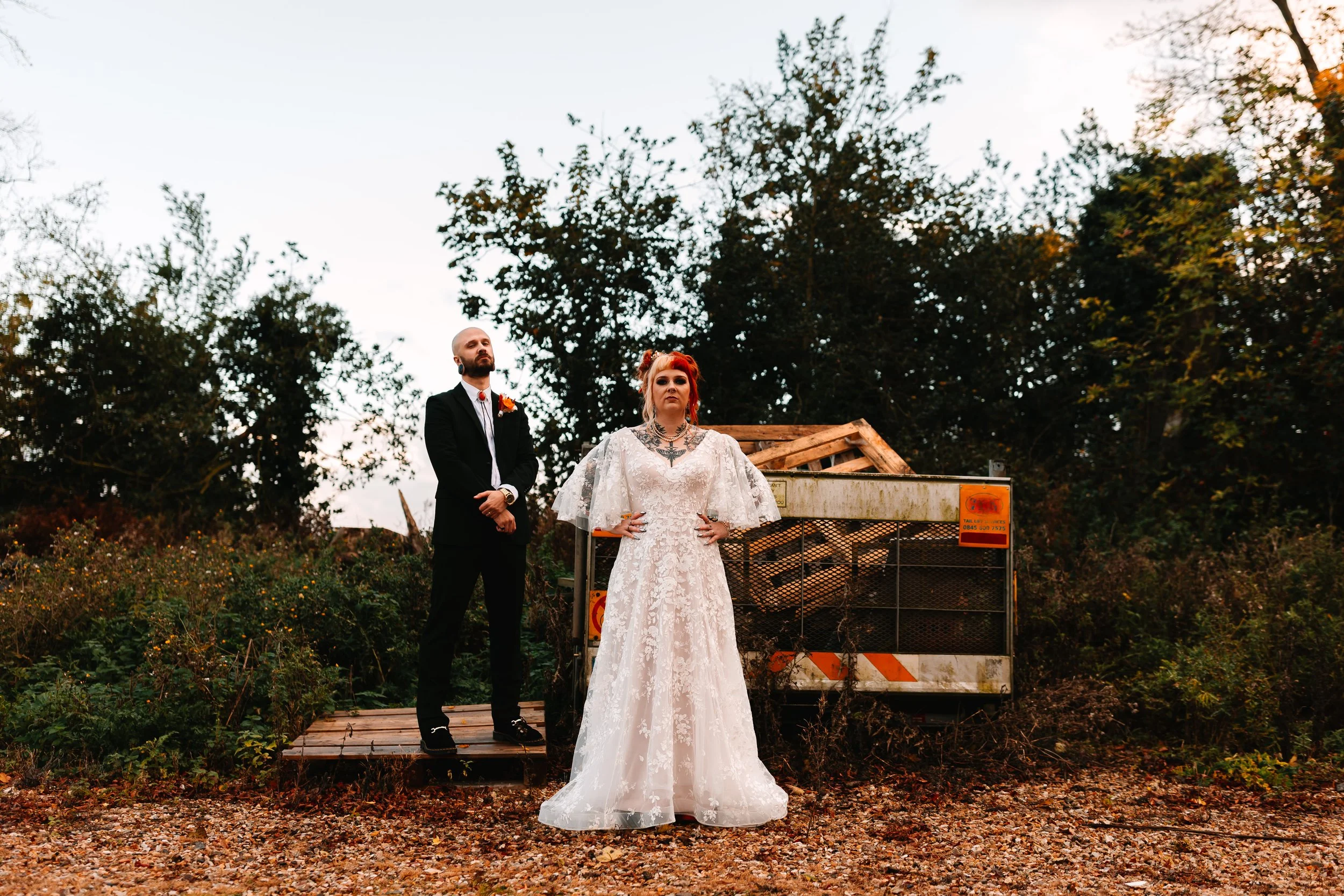 A bride & groom stand in a gritty, industrial outdoor spot, posing in front of a weathered trailer surrounded by overgrown plants. The bride’s lace dress contrasts with the rugged setting, while the groom stands slightly behind her in a black suit. T