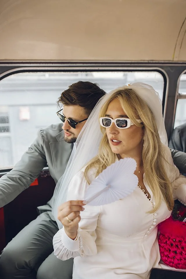 A couple dressed in wedding attire wearing sunglasses inside a vehicle, with the woman holding a paper fan.