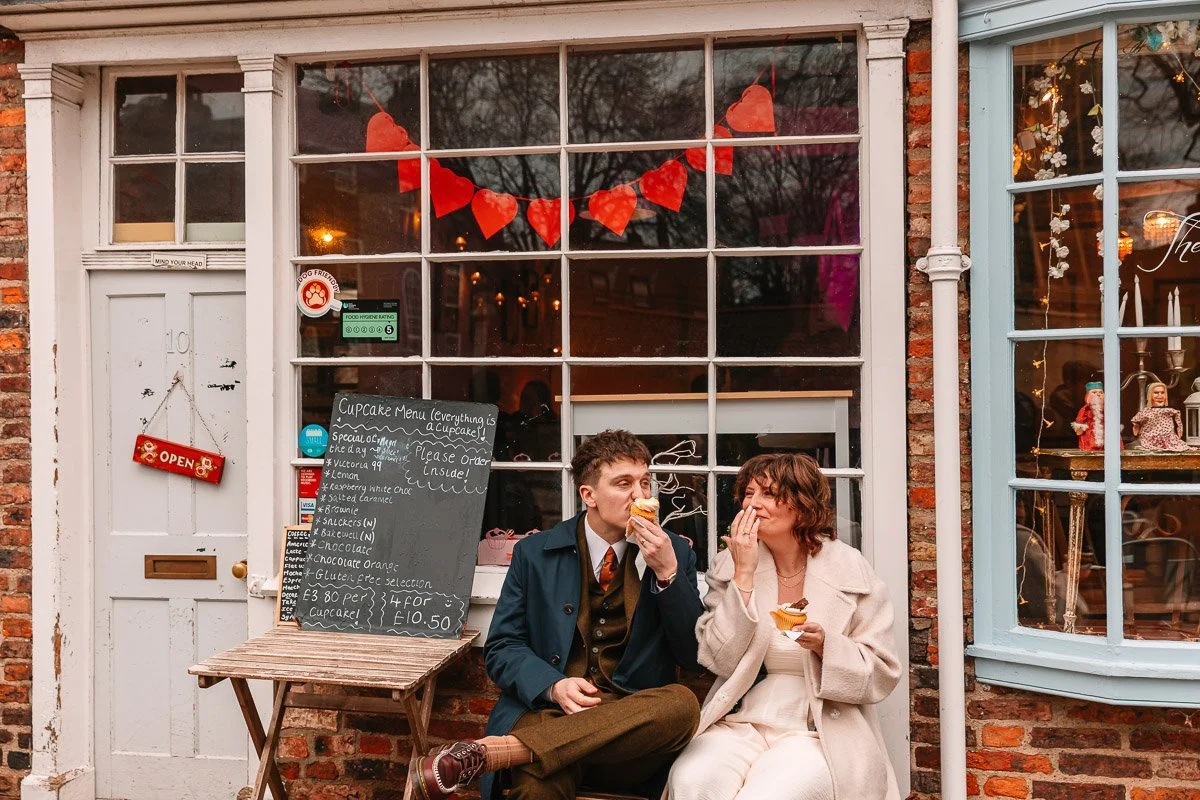 A Couple sit outside a cupcake shop in their wedding outfits eating cupcakes