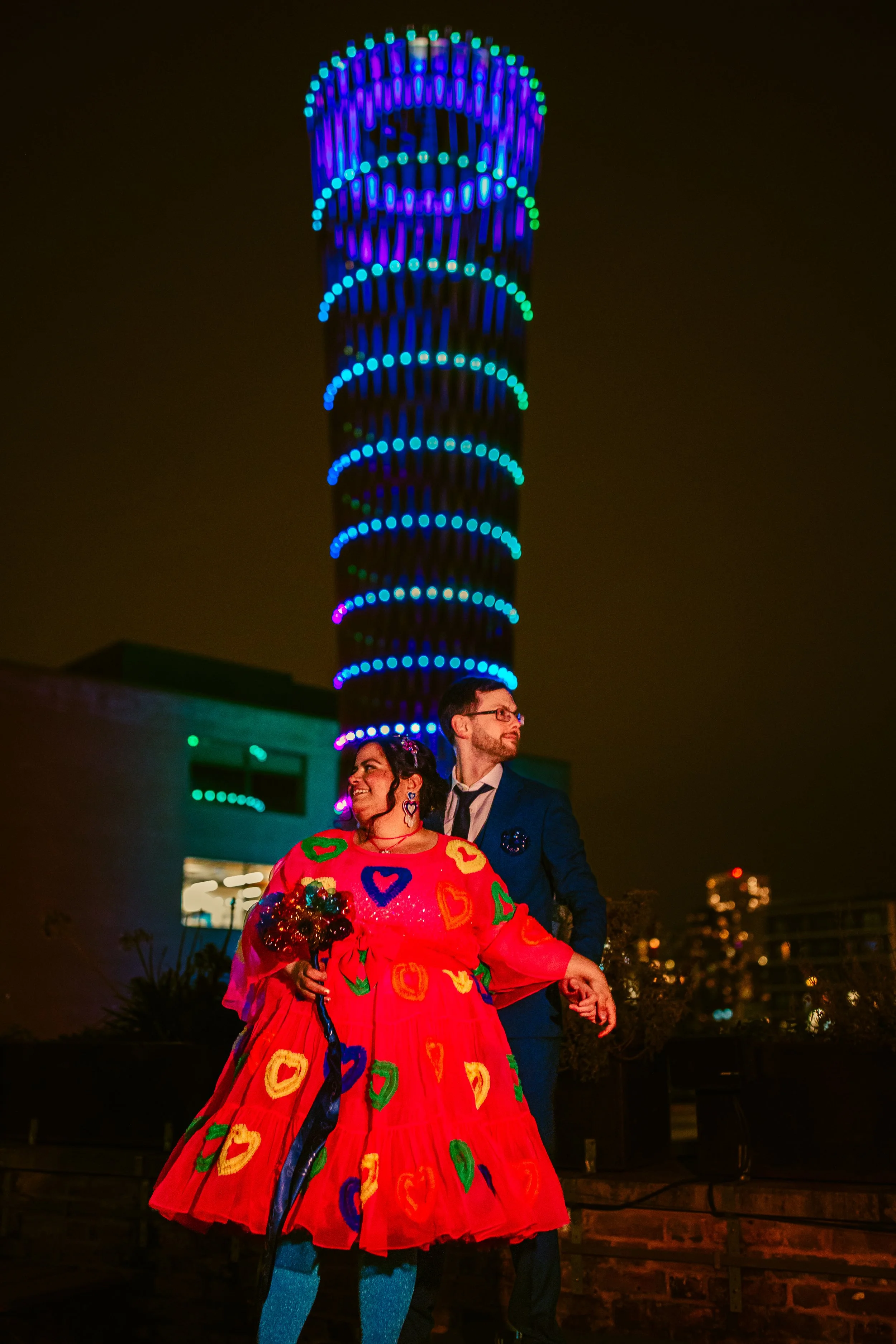 A joyful couple stands together at night on an outdoor rooftop with city buildings and colorful lighted towers in the background. The woman is wearing a bright, multicolored, patterned dress and holding a bouquet of flowers, while the man is in a dark jacket and glasses, smiling at her.